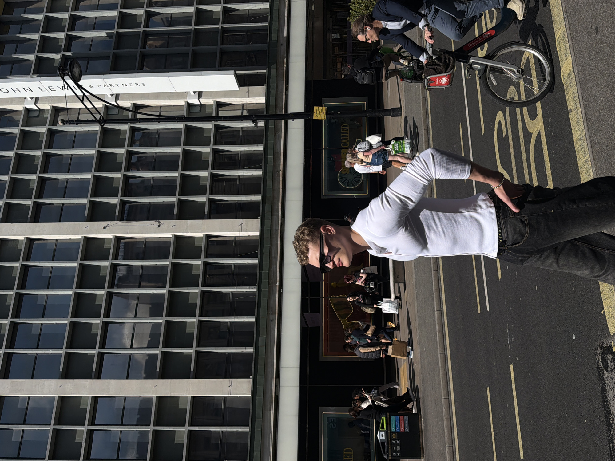 The image depicts a street scene in London, United Kingdom, adjacent to a building labeled "John Lewis & Partners." The building has a grid-like facade with numerous windows. A man wearing a white long-sleeved shirt, dark jeans, and sunglasses stands on the sidewalk. A person on a Santander bicycle rides in the opposite direction. Other pedestrians are present in front of the building. Yellow lines are painted on the asphalt street.