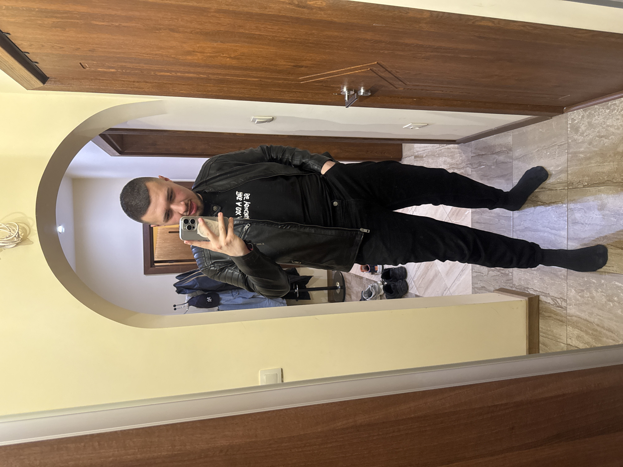 A man in a black t-shirt, black jeans, a black leather jacket, and black socks is taking a selfie in front of a mirror. The mirror is in a room with light yellow walls and a wooden door. There is a hallway visible through an arched doorway in the mirror's reflection. The hallway has light-colored tiled flooring and a coat rack with clothes on it. There are shoes on the floor near the coat rack. The location appears to be indoors, in a home or apartment. There is no information in the image to suggest this is in Sofia, Bulgaria.
