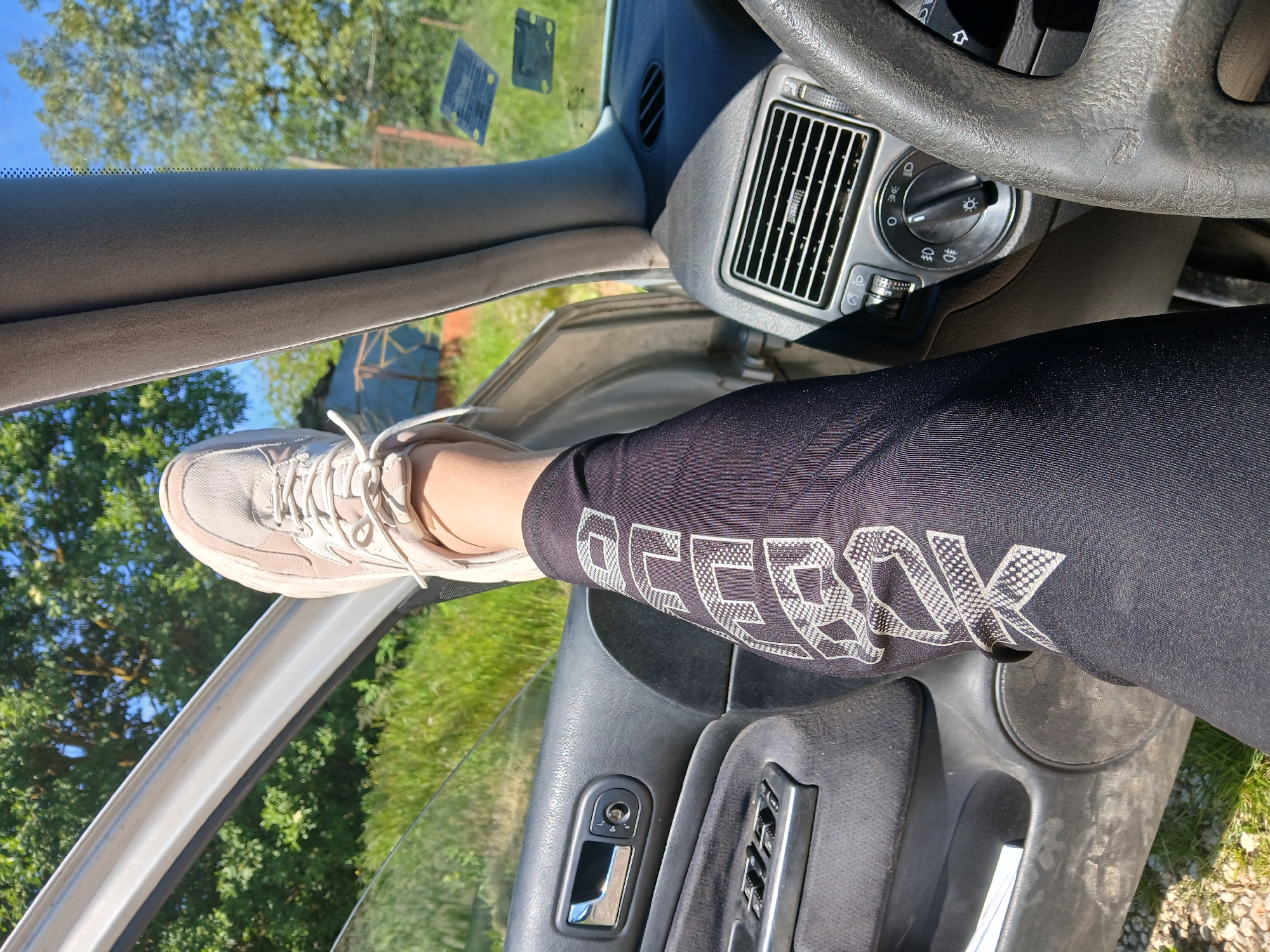 The image shows a person sitting in the driver's seat of a car. The person is wearing black Reebok leggings and a light-colored sneaker, which is placed on the open car door frame. The car's interior is visible, including the dashboard, steering wheel, air vents, and door controls. The outside environment is visible through the open door and windshield, showing trees and grass. There is no other information that specifies any location in the city of Sokolovo, Bulgaria.