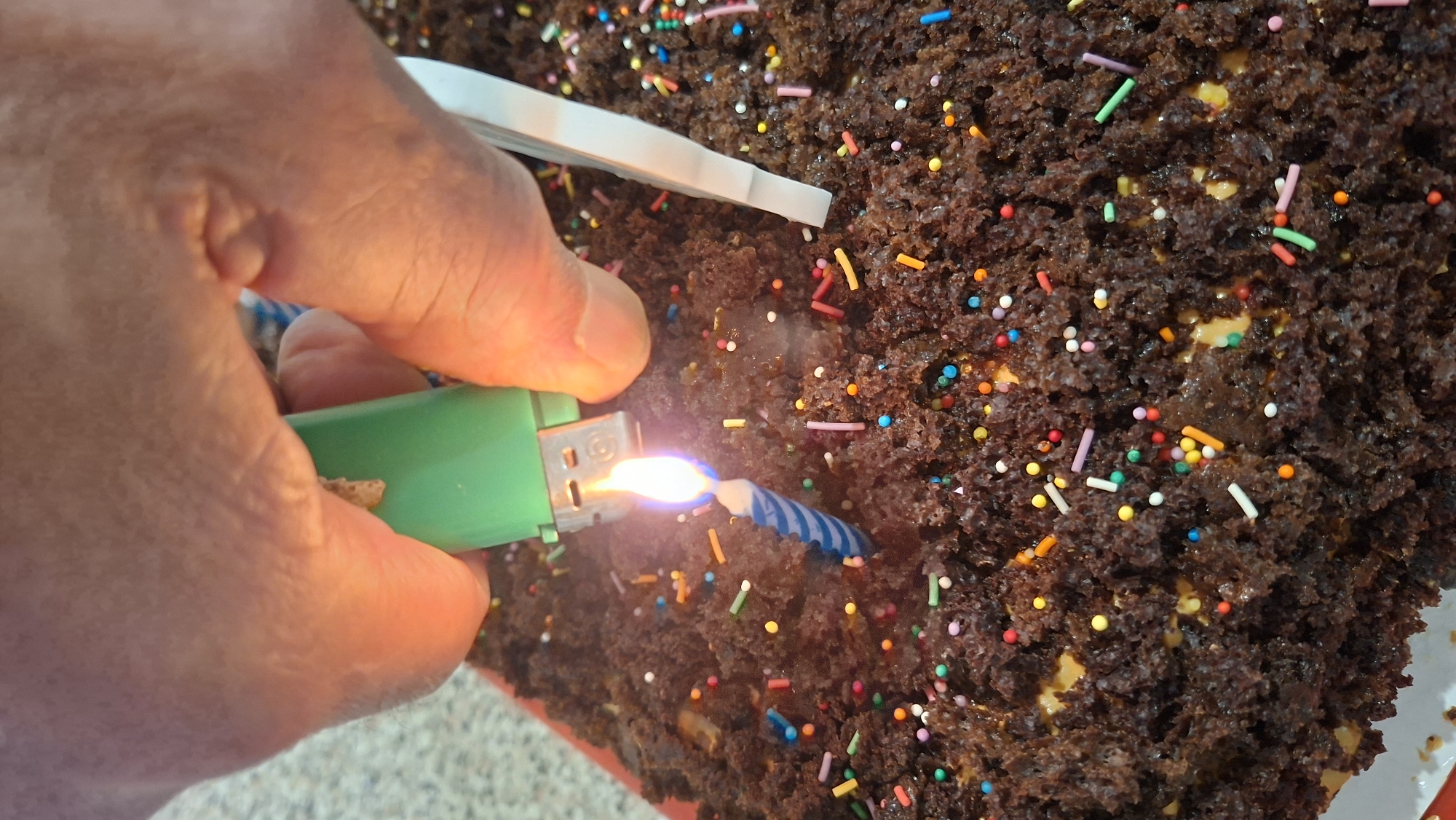 A close-up, top-down view captures a hand holding a green lighter, igniting a blue and white striped birthday candle on a dark chocolate cake. The cake is densely textured and adorned with colorful sprinkles in red, yellow, blue, green, pink, and white. A portion of a white plastic utensil is visible to the right of the candle. The background is a speckled beige surface, possibly a countertop. The lighting suggests an indoor setting, with the focus on the cake and the act of lighting the candle. There is no visible information about the city of Myadzyel, Belarus in this image.