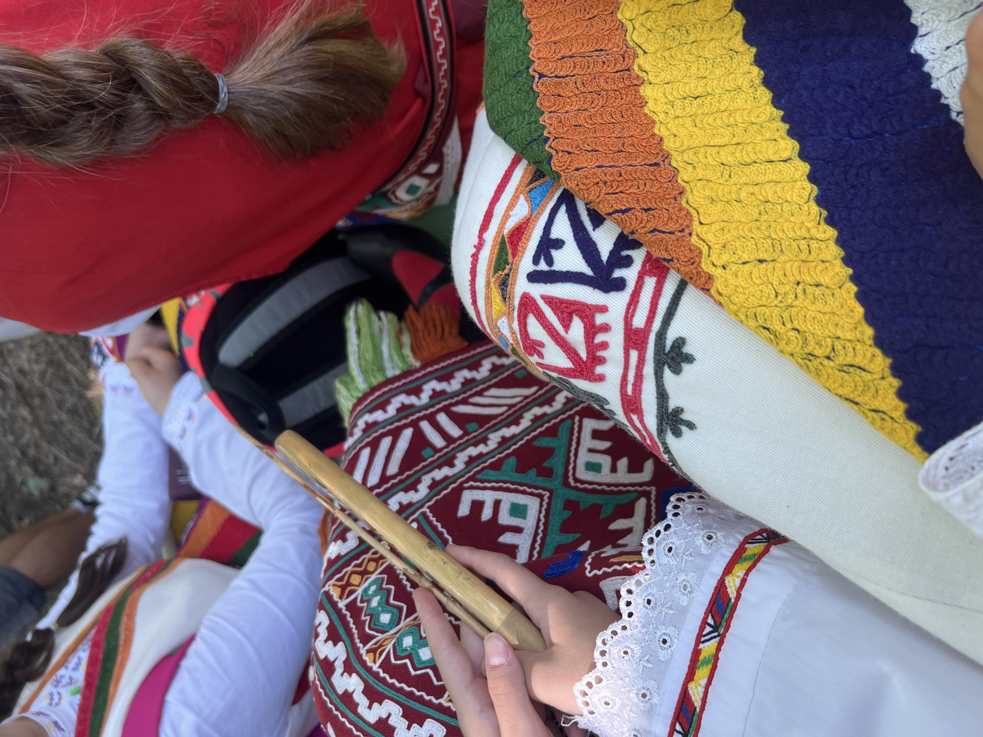 A close-up, high-angle view captures several individuals adorned in vibrant traditional Bulgarian folk attire during an outdoor gathering in Bolyarovo, Bulgaria. In the left foreground, a person's hands are clasped around a light-colored wooden wind instrument, resembling a kaval, which rests across their lap. This individual wears a white blouse with intricate eyelet lace trim on the sleeve cuff, below which is a band of colorful geometric embroidery in red, yellow, and green. Their lap is covered by a richly embroidered maroon or deep red fabric displaying prominent white and green stylized geometric patterns. To the right, another person is visible, wearing a plain red garment, from which a thick, dark brown braided ponytail, secured with a light gray hair tie, extends downwards. Adjacent to this individual and partially overlapping the embroidered fabric is a textured, horizontally striped woven item in bold bands of green, orange, yellow, and dark blue. In the upper left background, parts of another person's white garment with subtle embroidery are visible. The ground beneath the participants is indistinct, suggesting a natural, earthy surface under dappled light.