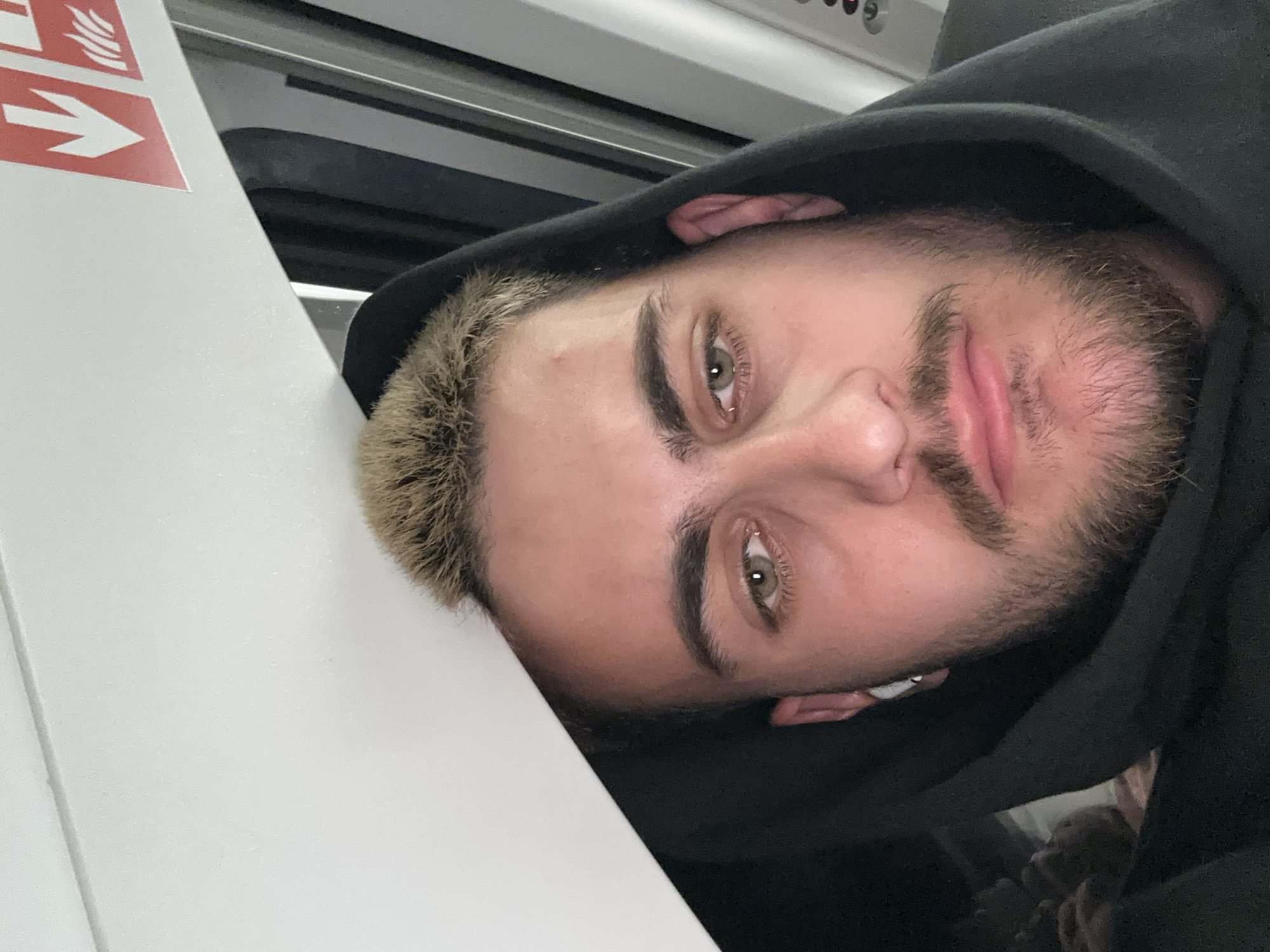 A close-up, rotated portrait shows a young man with striking green/hazel eyes, looking directly at the camera. He has short, light-blonde hair on top and darker stubble on his chin and cheeks. He is wearing a black hoodie, which frames his face, and a white earbud is visible in his left ear. The man appears to be resting his head against a light-colored surface, possibly a seat or wall.
The background suggests an interior setting, likely public transport such as a train or tram, given the overhead paneling and a dark, possibly windowed area behind him. To the left, a white panel features a red sign with a white arrow pointing right, accompanied by a symbol resembling stylized flames or smoke. This is indicative of a safety or emergency exit instruction, commonly found in transport vehicles. The overall lighting is soft but clear, suggesting it could be daytime inside the vehicle. The man's expression is neutral, bordering on pensive. Given the context, the setting is likely within a train or tram in Leverkusen, Germany.