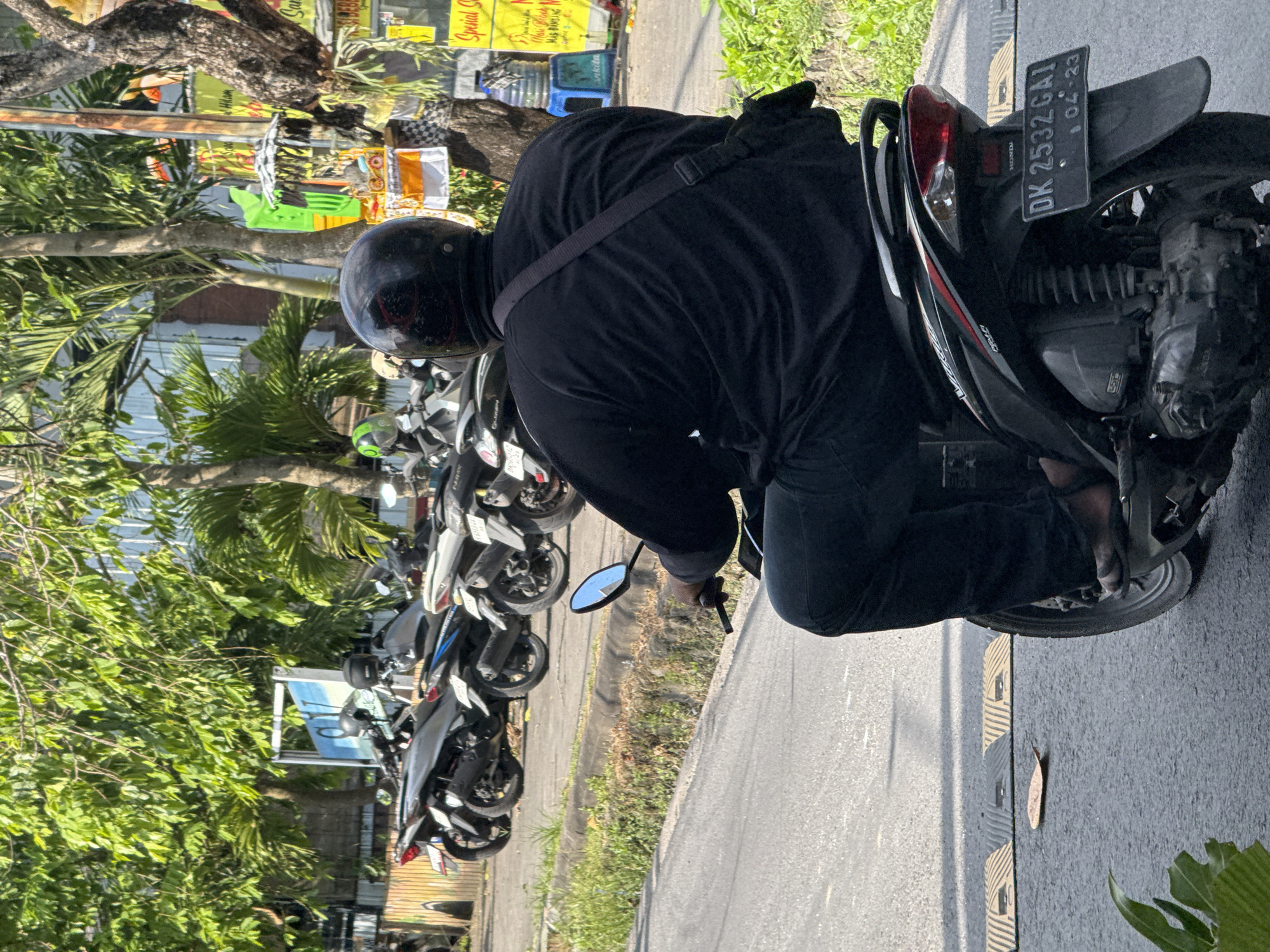 The image depicts a person wearing a black helmet and all-black clothing, seated on a parked black and silver Honda motorcycle. The motorcycle has a license plate reading "DK 2532 GAI" and an expiration date of "04-23". The person is positioned on a paved road with speed bumps visible. In the background, several other motorcycles are parked along a curb bordered by green foliage and trees, suggesting an urban or suburban street. The location is North Kuta, Indonesia, based on the motorcycle license plate.