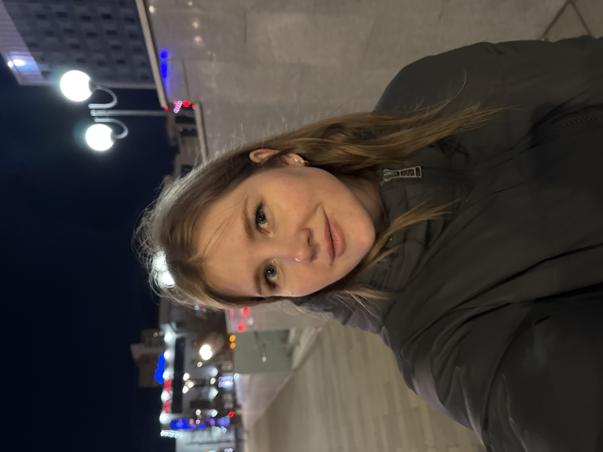 A medium shot depicts a young woman with light skin, blue eyes, and light brown hair, standing on a city street at night. She is wearing a dark puffy jacket. The street is lined with buildings, some of which are adorned with illuminated decorations. Two globe streetlights are visible on the left side of the frame. The photo was taken in Perm, Russia.