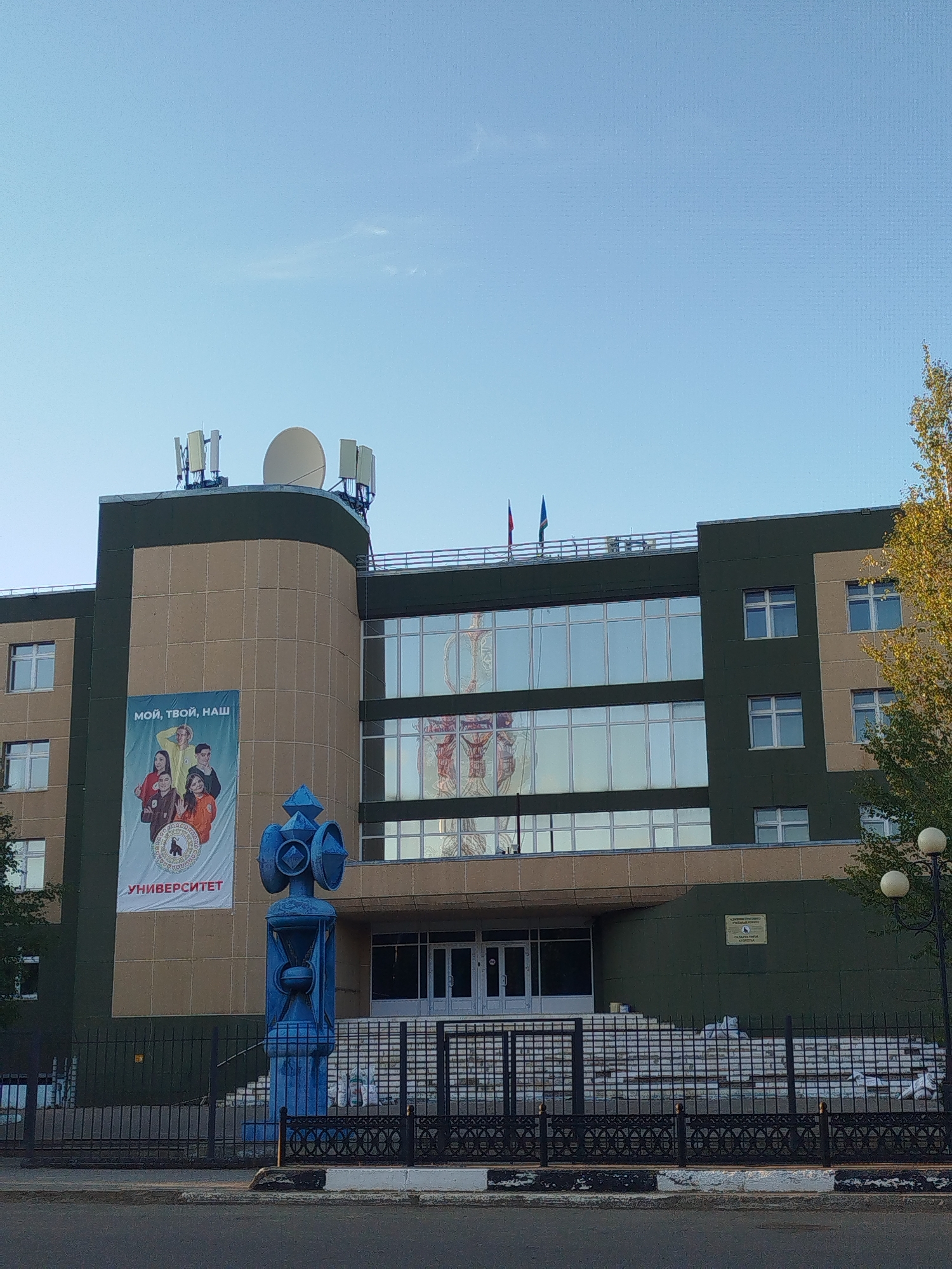 This is a frontal, eye-level photograph of the exterior of a university building in Mirny, Russia. The building is multi-story and features a modern, angular design with sections of tan and dark green cladding. Large, reflective windows dominate the upper floors. Two flags, one red and one green, fly from poles on the roof. A large banner is displayed on the front of the building, depicting four young people and text in Russian. To the left of the entrance, a vibrant blue, abstract sculpture stands on a pedestal. A black metal fence with decorative elements encloses the property. The sky is clear and blue, with a few wispy clouds. A deciduous tree with yellowing leaves is visible on the right side of the frame, and a lamppost with two globes stands nearby. The foreground shows a paved road with a curb.