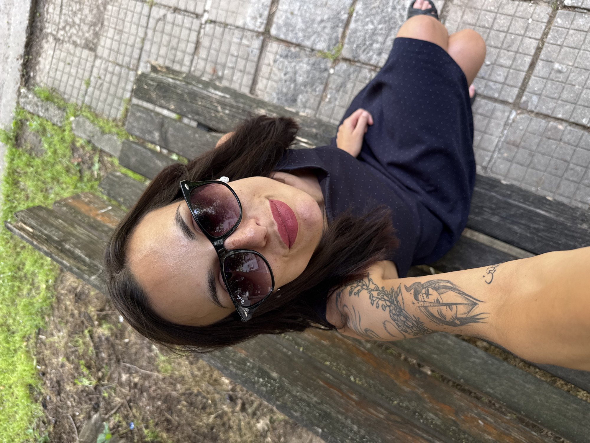 A medium-angle shot depicts a woman wearing sunglasses and a navy blue dress with small white polka dots, seated on a wooden park bench. Her legs are crossed at the knee, and she is looking up into the camera. Her left arm, visible in the lower right corner, features multiple tattoos. The bench is aged and weathered. In the background, there are both paved areas composed of small gray rectangular stones and patches of grass and dirt. While the precise location within the city of Pleven is not visually determinable, the setting appears to be an outdoor public space such as a park or a street.