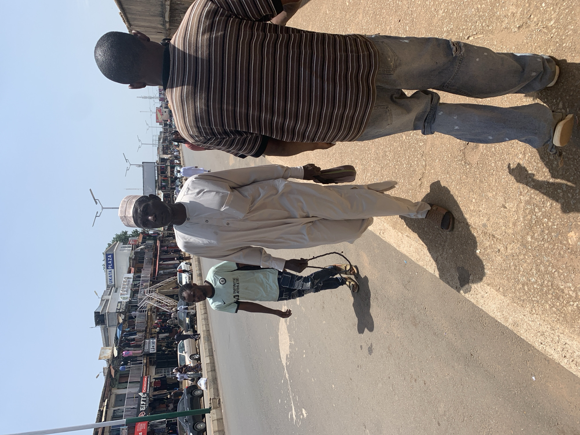 The image captures a street scene in the city of Jos, Nigeria, under a bright, clear sky. In the foreground, three individuals are walking on the right side of a road. The person closest to the viewer is seen from behind, wearing a horizontally striped t-shirt in shades of brown, white, and black, and light blue jeans. To their left, a man in a light-colored traditional robe and cap walks forward, carrying a small brown bag in his right hand. He is also holding a dark string of prayer beads. Further to the left, a younger man in a light blue t-shirt with white text and ripped blue jeans walks ahead of the other two, also carrying prayer beads in his hand.
The road appears to be a busy thoroughfare, with several cars visible. On the left side of the road, a row of shops and market stalls lines the street, with clothing and other goods displayed. A building with "UKATA PLAZA" signage is prominent in the background, indicating a commercial hub. The street is paved, with a dirt shoulder to the right where the people are walking. The overall impression is of a bustling urban environment with active commerce and pedestrian traffic.