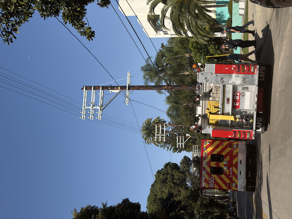 An image shows a firetruck and ambulance in a city street. Three firefighters stand near the back of the firetruck. The firetruck is numbered "E-82" and "LAFD." Utility poles stretch vertically with wires. A clear blue sky is above. Trees and buildings are also present.