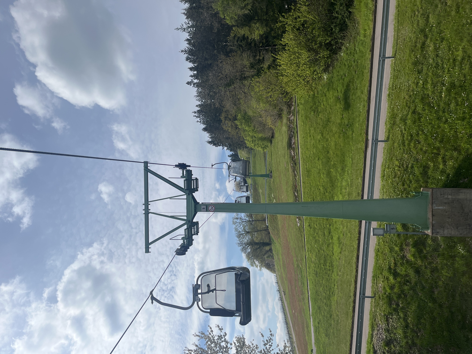 The image depicts a ski lift in Saarburg, Germany, viewed from a low angle. The sky is blue with scattered clouds, and the landscape is green with trees and grassy areas. A green metal ski lift pylon and the attached chair are prominent in the foreground. A road with a guardrail runs parallel to the lift line. Other ski lift pylons and chairs are visible in the distance.