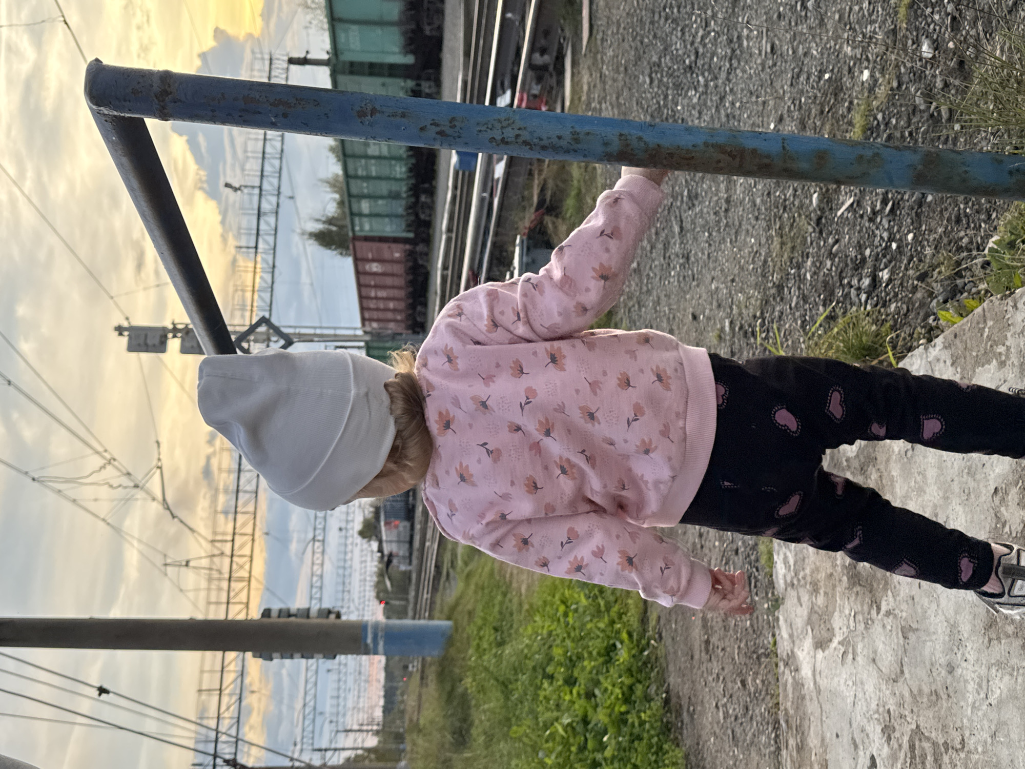 The image shows a young child from behind, wearing a white hat, a pink sweater with a floral pattern, and black pants with heart prints. The child is standing near a blue metal pole and appears to be looking towards a railway track. In the background, there are train cars and overhead electrical lines, suggesting the location is a train station or railway yard. The sky is partly cloudy with a warm sunset glow. The ground is covered in gravel and some grass. The child is positioned on what appears to be a concrete platform. While the image does not explicitly state the location, the query suggests the city of Komarihinsky, Russia.