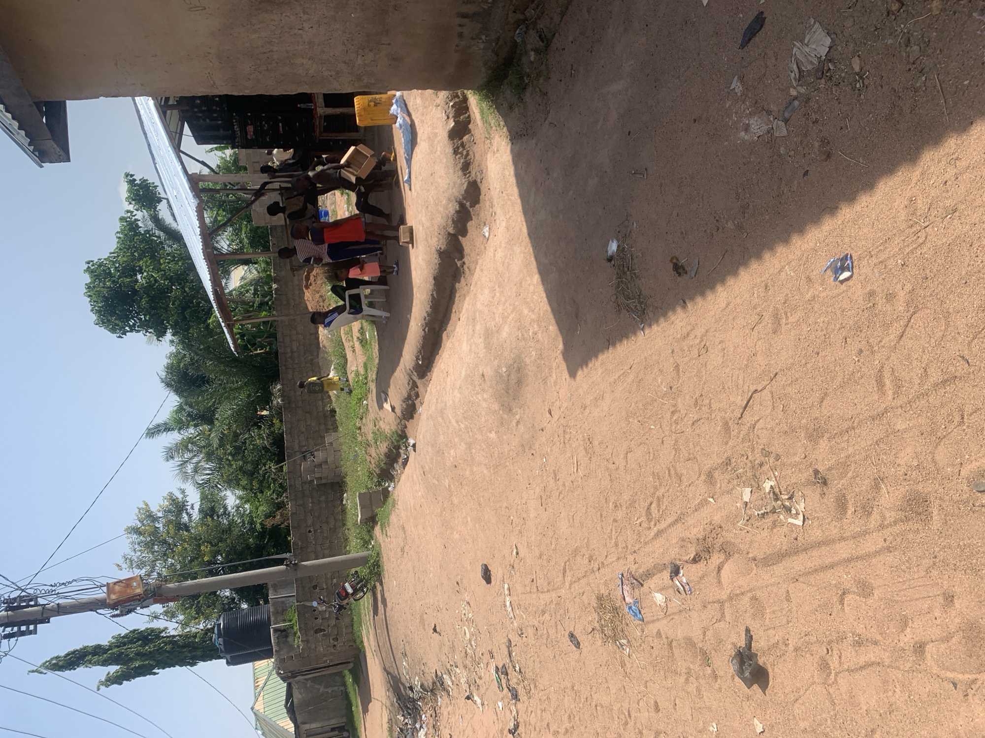 This media file captures an outdoor scene in Jalingo, Nigeria, likely during the daytime under a clear sky. The foreground is a dusty, sandy path with scattered debris including plastic bags, paper scraps, and dried vegetation, suggesting a somewhat unkempt environment. Tire tracks are visible in the sand, indicating vehicle movement.
In the mid-ground, a group of people, mostly children and young adults, are gathered. Some are dressed in casual clothing, including a child in a red shirt and another in a blue and white striped shirt. They appear to be engaged in some activity near a makeshift structure with a corrugated metal roof supported by wooden beams. Wooden crates are visible, and one person is holding a wooden plank or board. A yellow plastic container is also present. The group seems to be working or interacting, though the specific nature of their activity is unclear.
To the left, a brick wall and lush green trees provide a contrast to the sandy path. A utility pole with numerous wires and what appears to be an electrical transformer is a prominent feature, with a large black water tank situated nearby. A motorcycle is parked against the brick wall. In the distance, more trees and buildings can be seen. The overall impression is of a community setting, possibly a residential area or a workspace, with evidence of daily life and ongoing activities. The lighting suggests it is daytime, with sunlight casting shadows across the path. There is no visible text in the image.