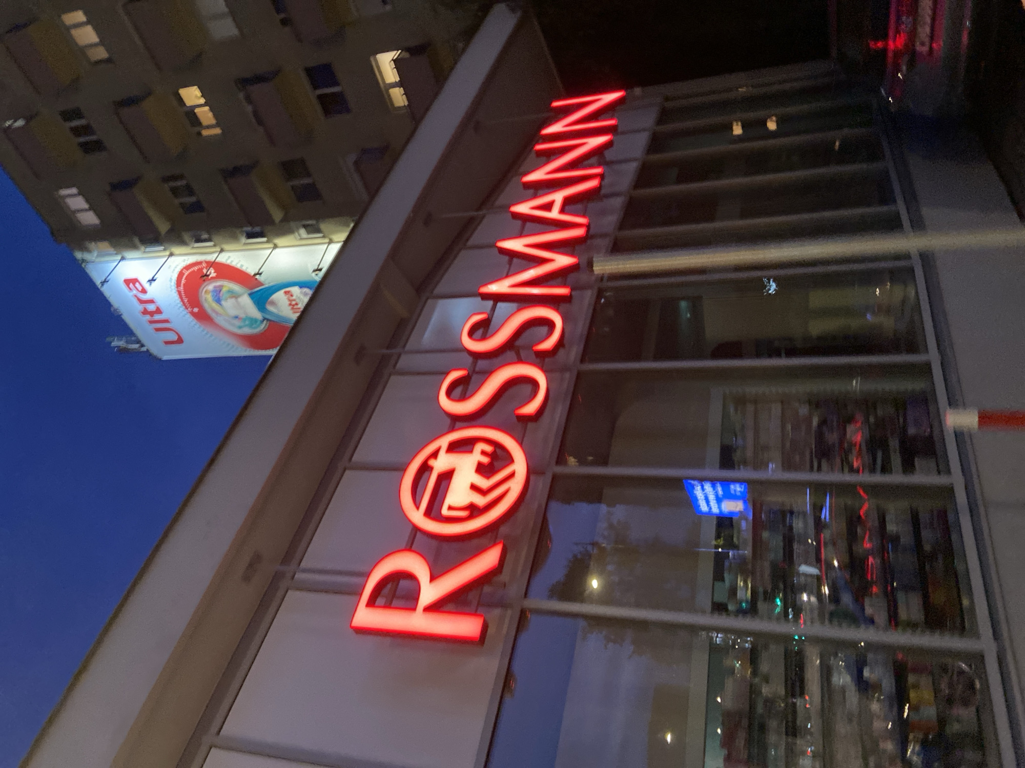 The image shows a Rossmann store in Warsaw, Poland, at dusk or night. The store's prominent red neon sign, "Rossmann," is illuminated and visible across the facade. The building appears to be a commercial property with large glass windows reflecting the interior of the store and the dark blue sky. Adjacent to the Rossmann building is a multi-story apartment building with illuminated windows. A large billboard advertising "Ultra" detergent is mounted on the side of the apartment building, also illuminated. The scene suggests an urban environment with commercial and residential buildings.