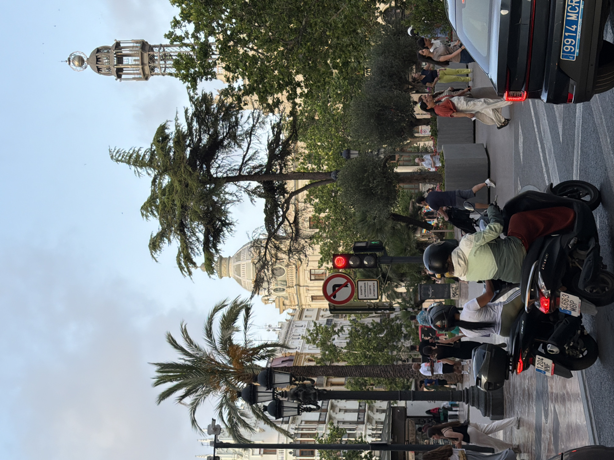 In the city of Unknown, Unknown, on a paved street, a group of people are seen with buildings in the background. In the foreground, two scooters are parked, one black and one dark colored. The black scooter has a license plate that reads "4663 MBP". The other scooter has a license plate that reads "1204 NMF". On the right side of the image, a black car is parked, with its taillight illuminated. The car's license plate reads "9914 MCR". A traffic light is visible, showing a red signal. Behind the scooters, two individuals are seated on a scooter, both wearing helmets. One is wearing a white shirt and green pants, and the other is wearing a white shirt and dark pants. In the middle ground, several people are walking, and palm trees and other foliage are present. The background features buildings with architectural details and a tall, ornate tower. The sky is cloudy.