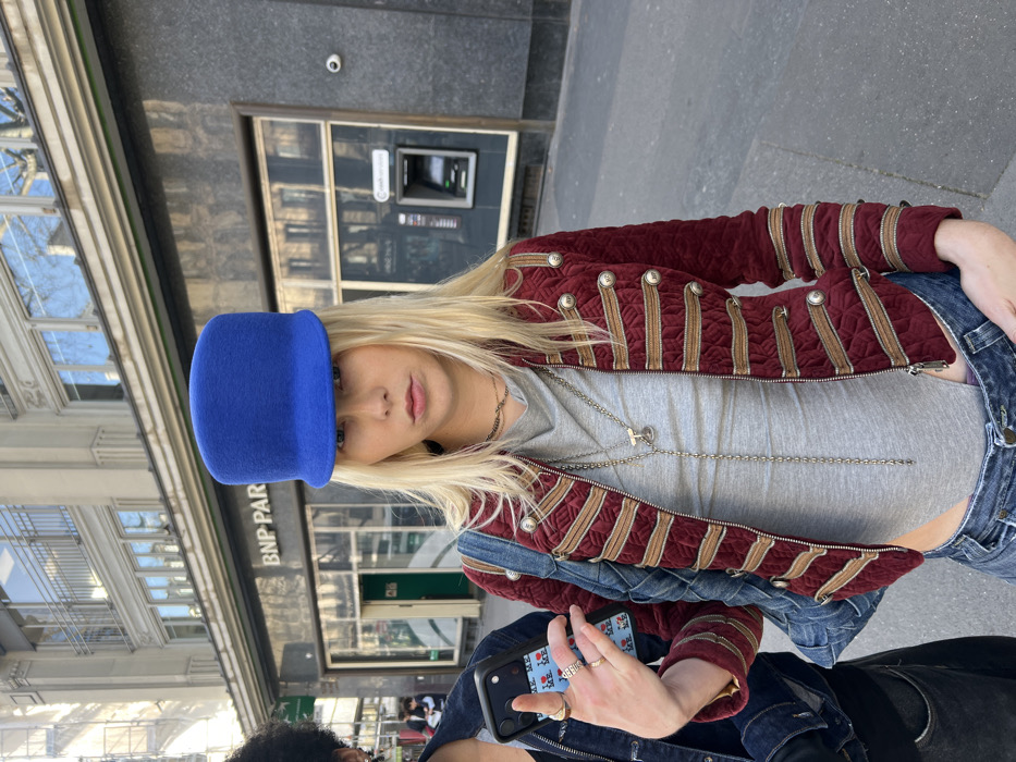 An outdoor, eye-level photograph captures two individuals on a Parisian sidewalk in front of a BNP Paribas bank branch. The central figure, a person with long blonde hair, wears a royal blue flat-top cap, a maroon quilted jacket with gold military-style braiding and silver buttons, a gray high-neck top, and blue denim jeans. This individual displays multiple rings on their left hand, which is held up, while two silver necklaces are visible. Partially visible to the left is a second individual with dark, curly hair, wearing a dark denim jacket and holding a black smartphone with a light blue case featuring "I LOVE NY" iconography. The background consists of a multi-story building facade with prominent "BNP PARIBAS" signage, reflective windows, and an automatic teller machine (ATM) on the right side. The architecture indicates a typical European urban environment, confirmed by the bank's presence in Paris, France. The scene is illuminated by natural daylight.