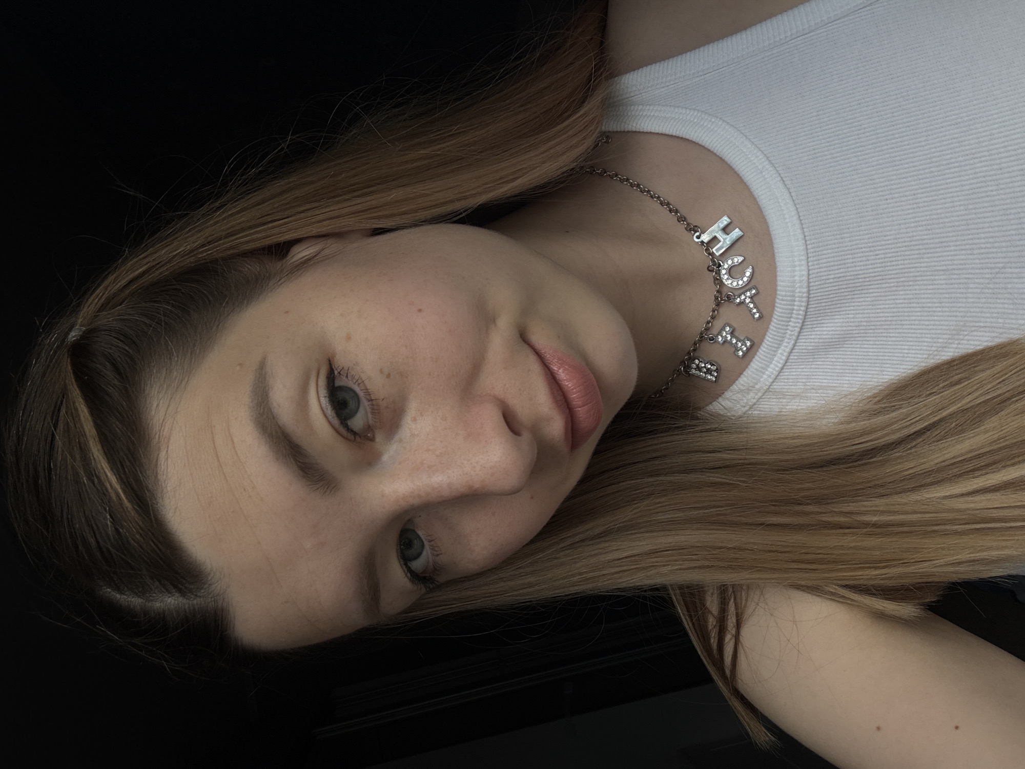 A portrait of a light-skinned woman with blue eyes and blonde hair wearing a white tank top and a silver necklace with the word "HOTCH" spelled out in sparkling letters. The background is black.