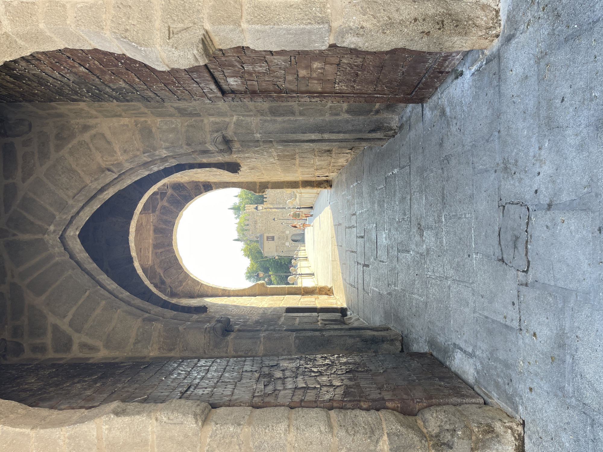 The image shows an arched passageway made of stone. The passageway is covered by multiple arches, with the furthest one revealing a bright, sunlit exterior. Two large, heavy wooden doors flank the sides of the entrance. The floor is paved with cobblestones. Two individuals, a man and a woman, are walking away from the camera on the right side of the passageway, in the distance. The scene is located on the San Martin Bridge in Toledo, Spain. The bridge dates back to the 14th century and connects the city to the San Martin Monastery.
