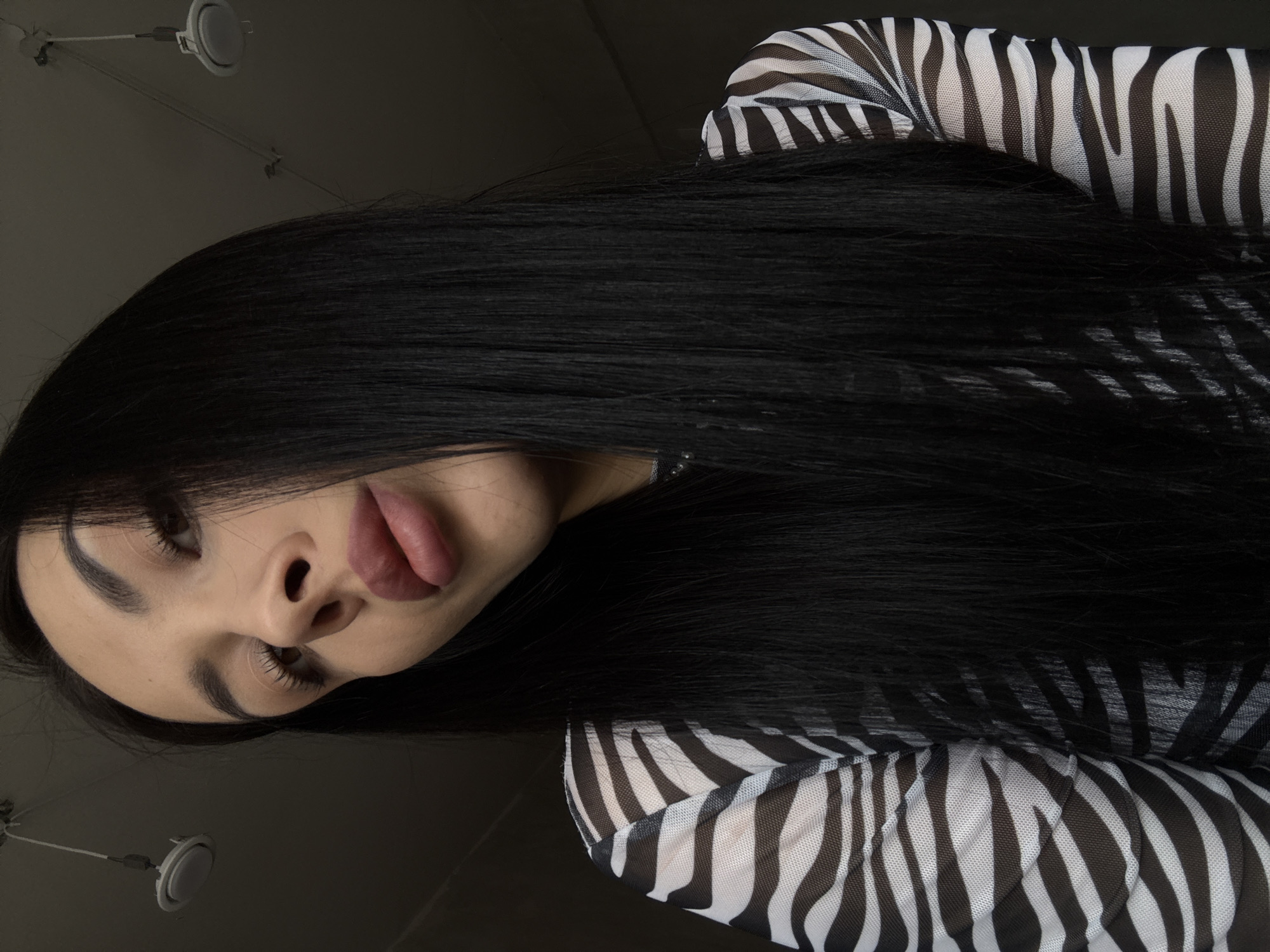 A woman with long, dark hair lies on her side wearing a zebra-striped, mesh top. The photo is taken from a low angle, and overhead lighting is visible in the background. The woman's face is partially obscured by her hair. Her lips are slightly parted. The location is indeterminable and cannot be identified as Votkinsk, Russia based on the provided image.