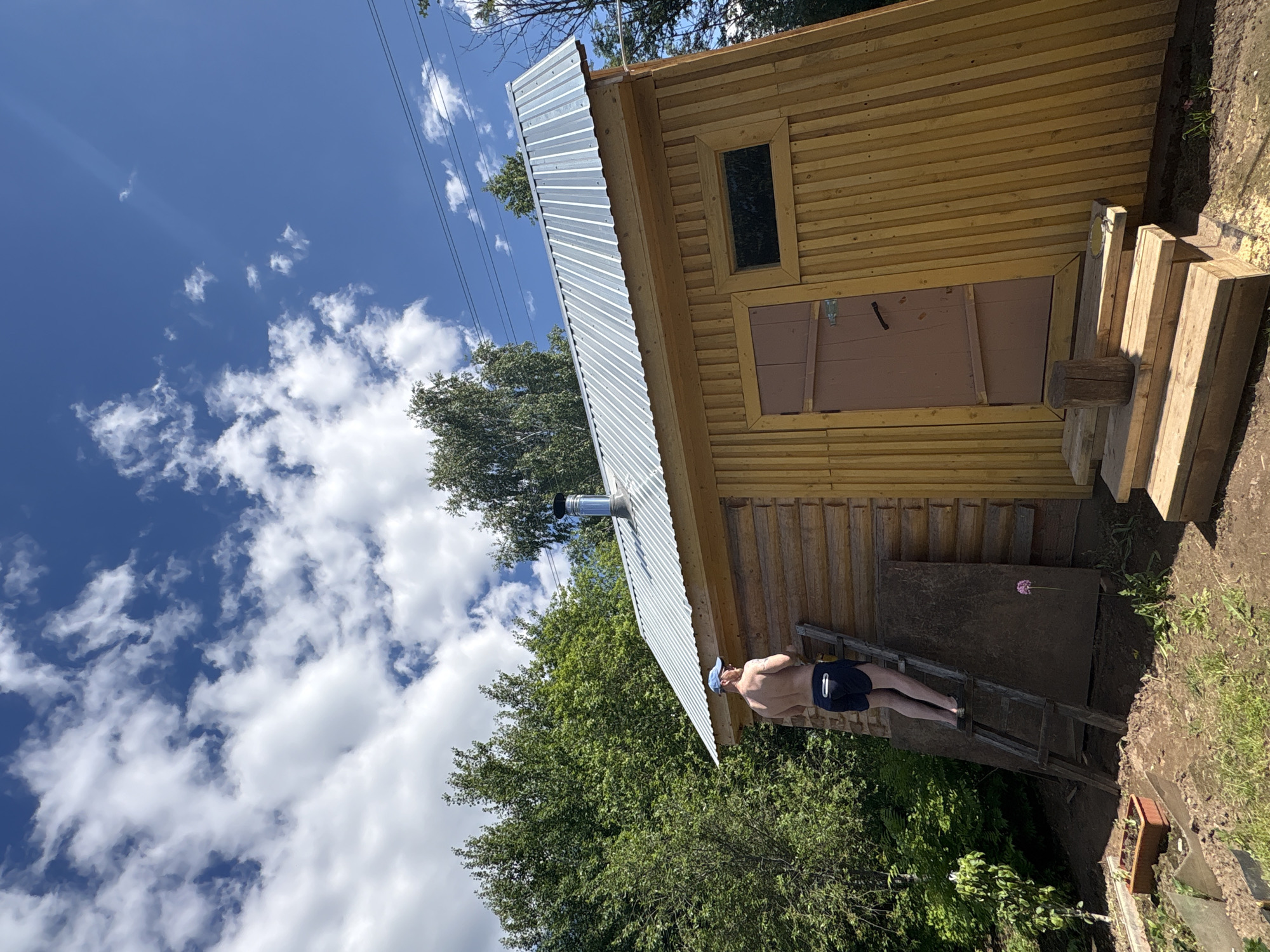 The image depicts an outdoor scene in Italmas, Russia, under a bright blue sky with scattered white cumulus clouds. An adult male, shirtless, wearing dark blue shorts with white trim and a light blue baseball cap, stands on a wooden A-frame ladder. His back is mostly to the viewer as he faces the upper portion of a rustic wooden building, engaged in an unseen task. Tattoos are visible on his upper back and right shoulder.
The building is a single-story structure with walls constructed from light-colored wood, appearing as logs on the left and vertical planks on the right. Its roof is made of corrugated galvanized metal, sloping upwards from left to right, and a shiny, cylindrical metal chimney pipe extends from it. The front of the building features a simple, light brown wooden door with a dark handle and a rectangular window with dark glass to its right. Three wooden steps lead up to the door, with a small wooden stump and a small reflective metal object resting on the top step.
The ground immediately surrounding the building is disturbed dirt with some patches of green grass. A dark, rectangular panel, possibly wood or fiberboard, leans against the building wall beneath the ladder. To the left of the ladder, a small garden area contains a brown planter. A single purple flower grows out of the dirt near the base of the ladder. Dense green deciduous trees frame the background, and thin black power lines are visible in the upper right quadrant of the sky.