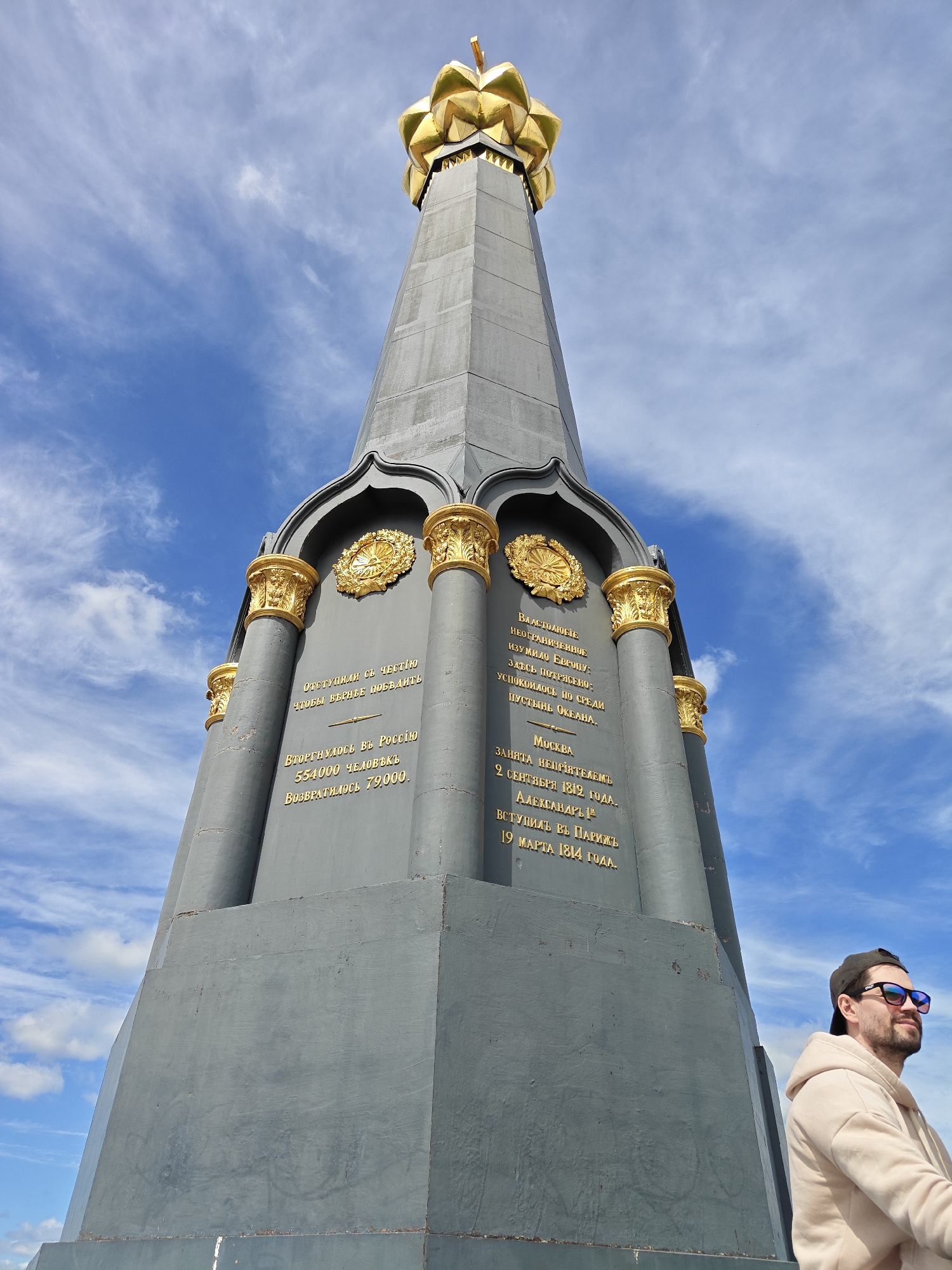 The image depicts a monument, presumably the Main Monument on the territory of the Borodino Field Museum-Reserve in Borodino, Russia. The monument is gray with gold detailing and inscriptions. It is an obelisk shape, tapering towards the top, which features a gold, lotus-like structure. The sky is blue with scattered white clouds. In the foreground on the right side of the image, a person wearing a hoodie, sunglasses, and a baseball cap is partially visible. The person is Caucasian, with a beard, and appears to be standing or leaning near the monument. The monument has vertical columns with decorative Corinthian-like capitals at the top and inscriptions with Russian text in gold lettering on each of its sides. The text appears to commemorate historical events, possibly the Battle of Borodino.