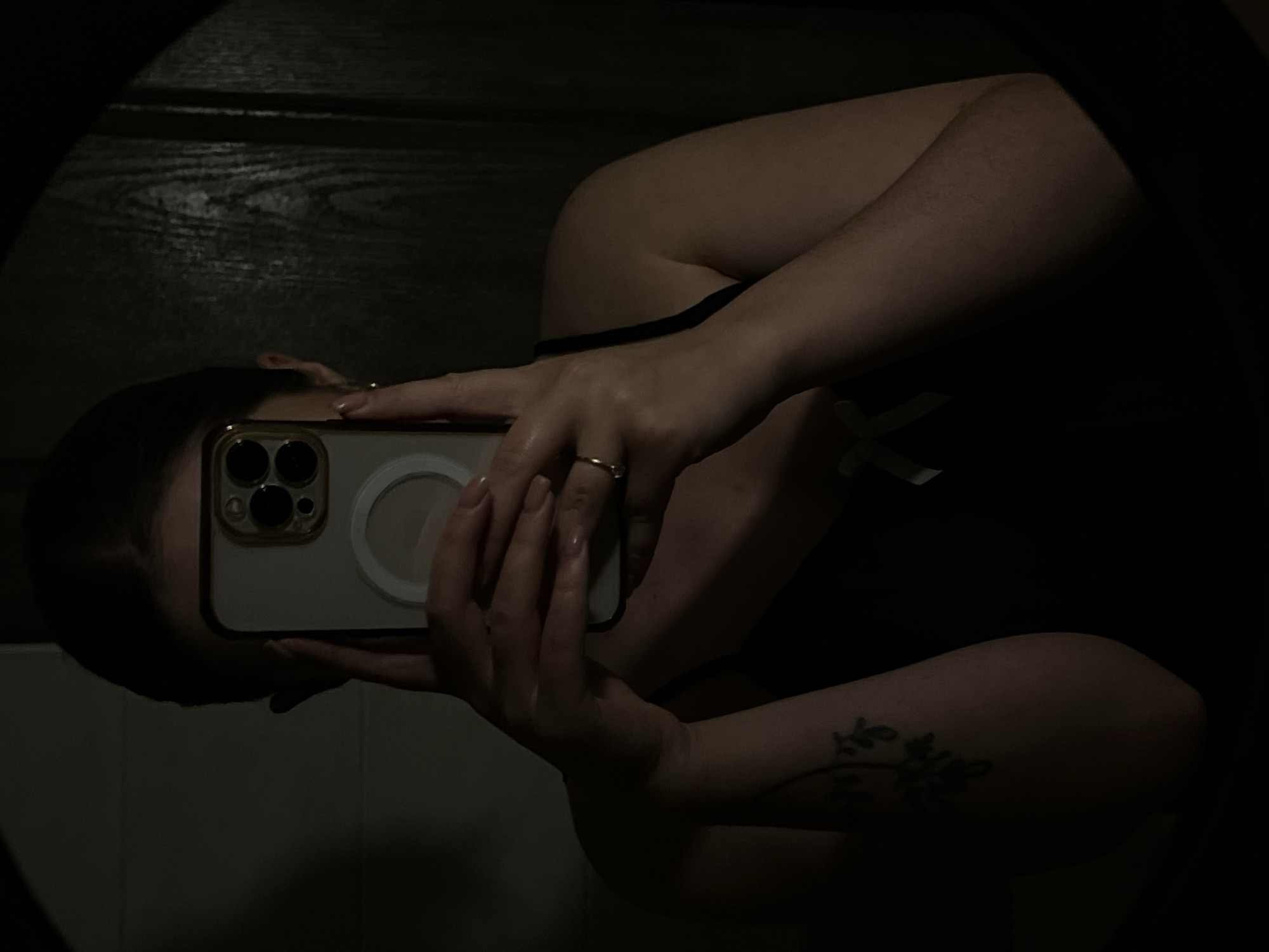 The image shows a person taking a selfie in a mirror. The person is holding a smartphone with a gold-trimmed case that has three camera lenses on the back. The person is wearing a ring on their finger. The background includes a dark wood-paneled wall and white-tiled wall. There is a small tattoo of leaves and flowers on the person's left forearm. There is no information suggesting this photo was taken in Ternopil, Ukraine.