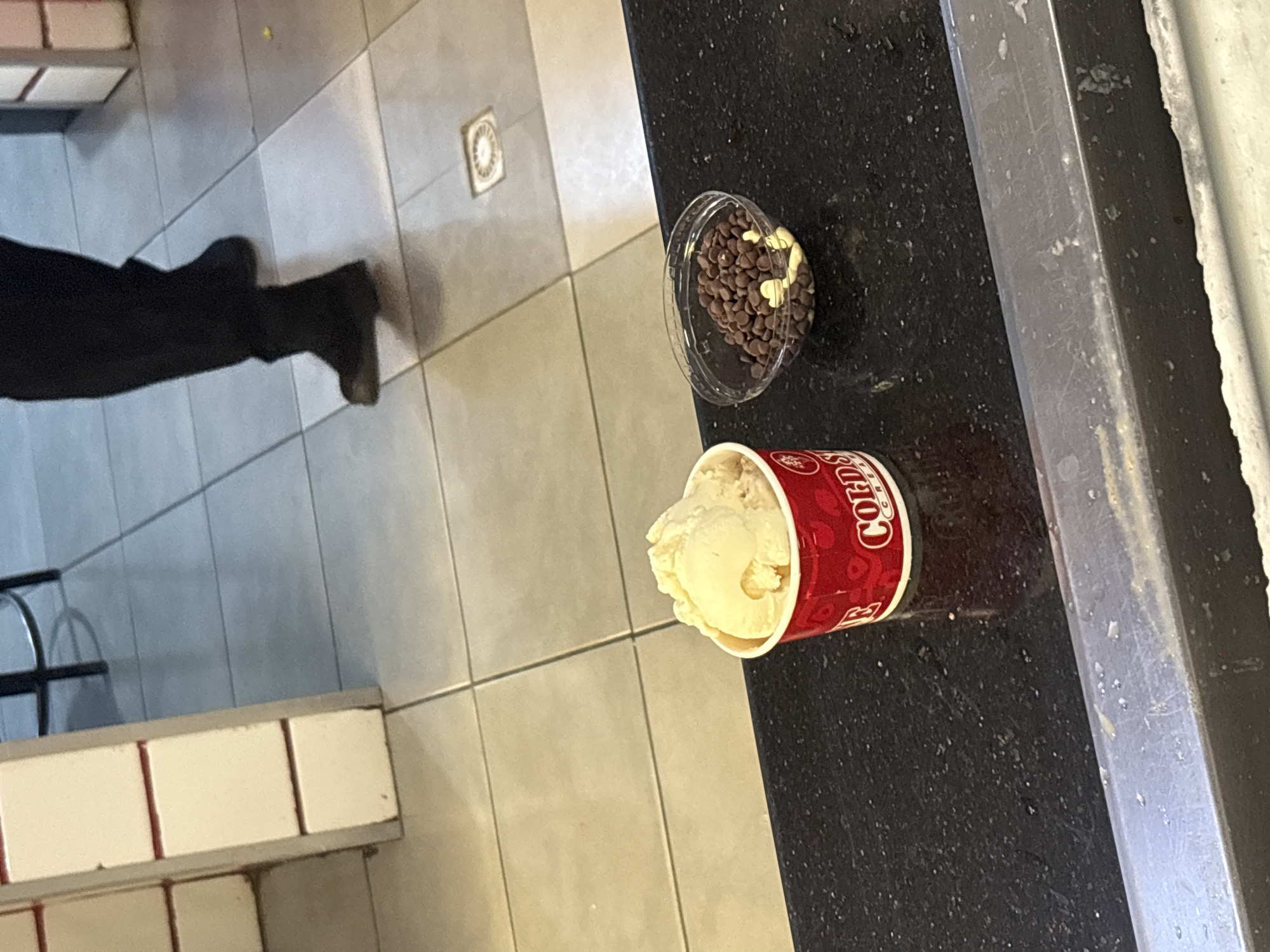 A partially eaten cup of Cold Stone Creamery ice cream sits on a black countertop. A small, clear plastic container holding chocolate chips and possibly some white chocolate chips is also on the counter. A person wearing dark pants and shoes is partially visible in the background. The scene is likely inside a Cold Stone Creamery store. It is not possible to determine if the store is located in Abuja, Nigeria from this image alone.