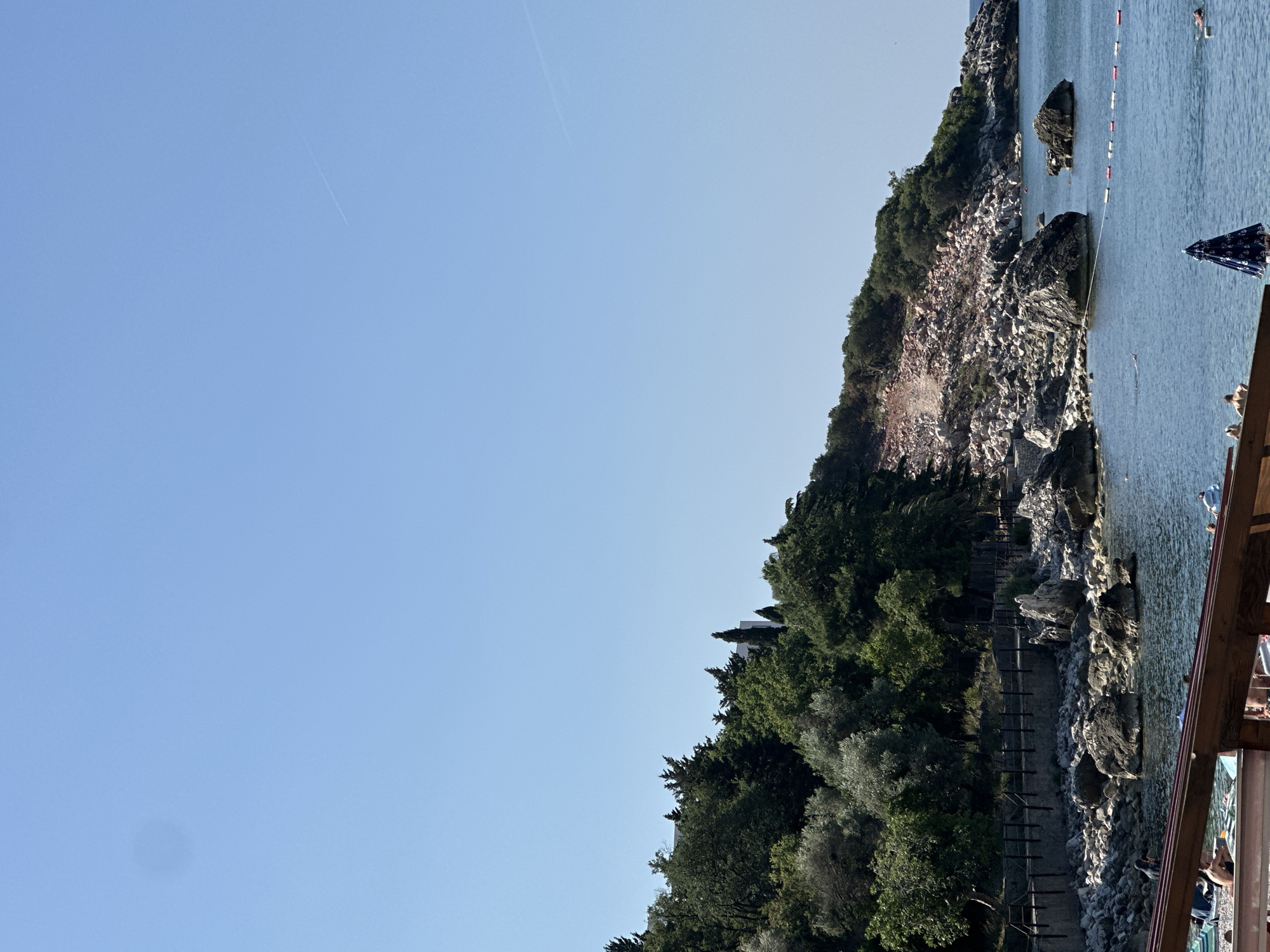 The image depicts a coastal scene in Boreti, Montenegro, on a clear, sunny day. The foreground shows a wooden structure with people visible on a beach below. The water is calm and blue, with several buoys marking a swimming area. To the left, a rocky shore leads to a hillside covered in lush green trees. The hillside slopes upwards, revealing a large pile of debris and exposed earth, suggesting recent construction or excavation. The sky is a vast expanse of light blue with a faint contrail from an airplane. Several rocks are visible in the water, some emerging as small islands. The overall impression is of a natural landscape impacted by human activity.