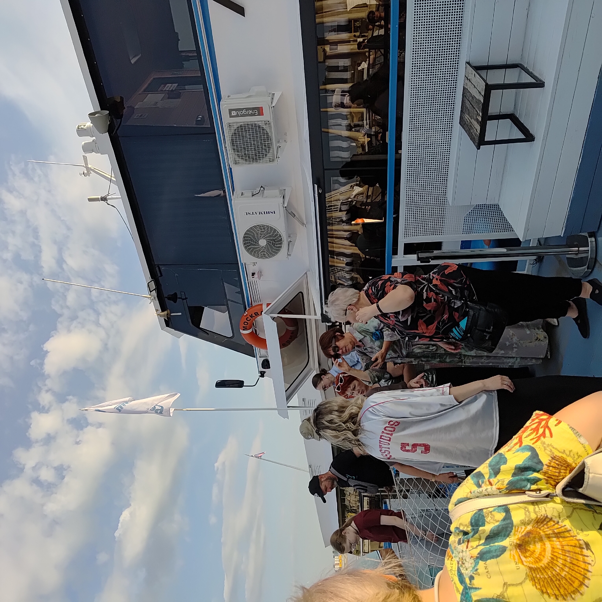 The image depicts a group of people on the deck of a boat, likely in Ufa, Russia. The boat appears to be a passenger vessel, with large windows indicating an enclosed cabin area. Several individuals are visible, dressed in casual attire. One woman in the foreground is wearing a bright yellow top with a map and seashell print. Behind her, a person in a grey t-shirt with "STUDIOS" and the number "5" printed on it is visible. Other individuals are standing and conversing, some looking at their phones.
The deck is made of a blue-colored material, and there is a white bench with a small wooden table on it. A white flag with blue lettering is fluttering in the wind from a mast. Two air conditioning units are mounted on the side of the boat's cabin. An orange life preserver with Cyrillic text is also visible. The sky above is cloudy with patches of blue. The overall scene suggests a leisurely outing on the water.