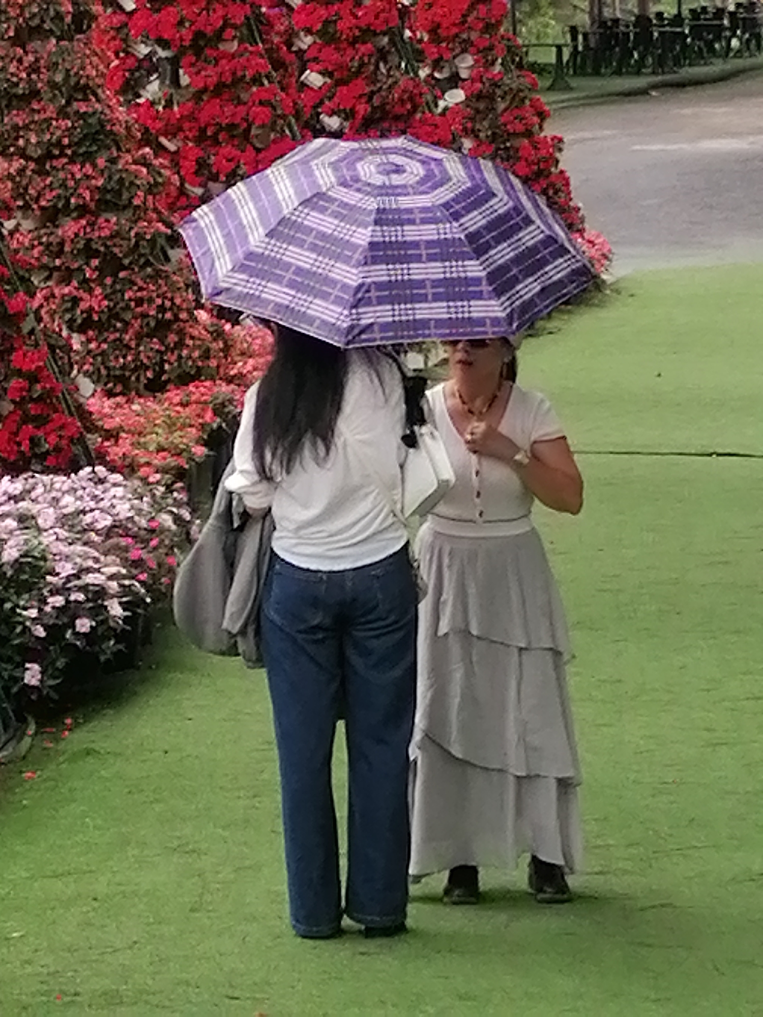 Two women stand under a purple and white checkered umbrella on a green lawn, with a dense backdrop of red and pink flowers. The woman on the left wears a white shirt, blue jeans, and holds a gray jacket, facing away from the camera. The woman on the right wears a light-colored top, a tiered gray skirt, sunglasses, and a necklace. The scene is set at a location with manicured green grass, likely a park or garden in the city of Da Lat, Vietnam. A paved path and benches are visible in the background.