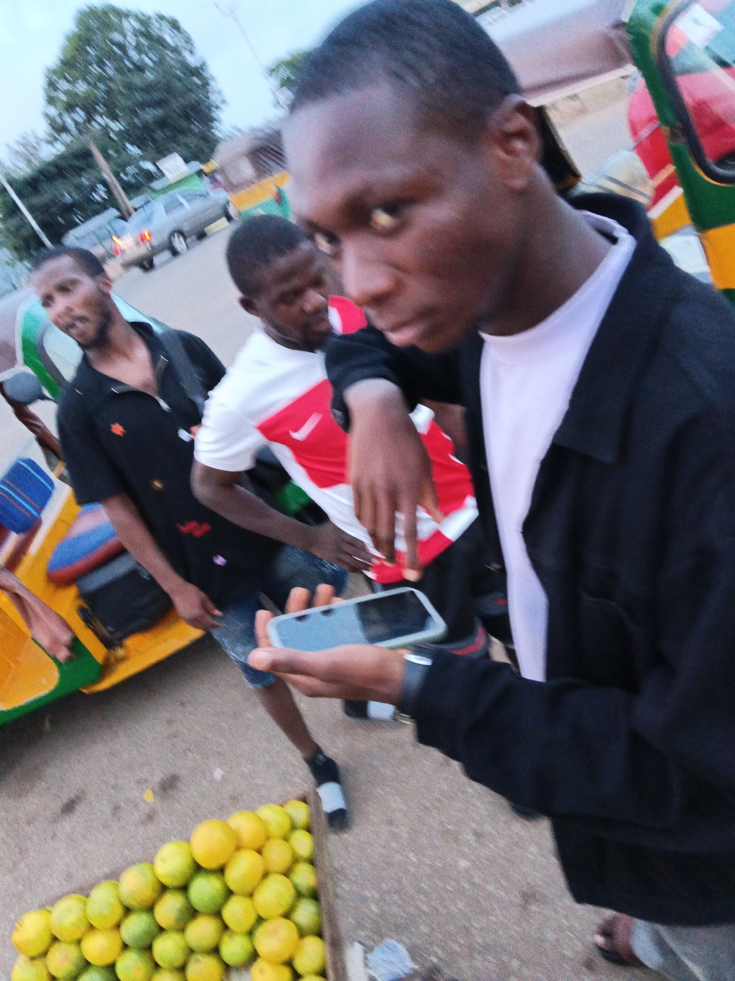 The image captures a street scene in Bukuru, Nigeria, with a focus on several individuals and a common mode of transport. In the foreground, a person is holding a smartphone, seemingly taking a selfie or recording a video, with their face partially visible. To their left, another individual wearing a black shirt with decorative elements stands near a yellow and green auto-rickshaw, also known as a "Keke Napep" in Nigeria. Behind them, another person in a white shirt with red detailing is visible, also near the auto-rickshaw. The auto-rickshaw itself is a prominent object, brightly colored and appearing to be in use. In the lower part of the image, a pile of oranges is neatly arranged, suggesting a roadside fruit vendor. The background shows elements of a typical urban environment, including a road, other vehicles (one appearing to be a gray sedan), and trees. The lighting suggests it is either early morning or late afternoon. The overall impression is a candid snapshot of everyday life in a Nigerian city.