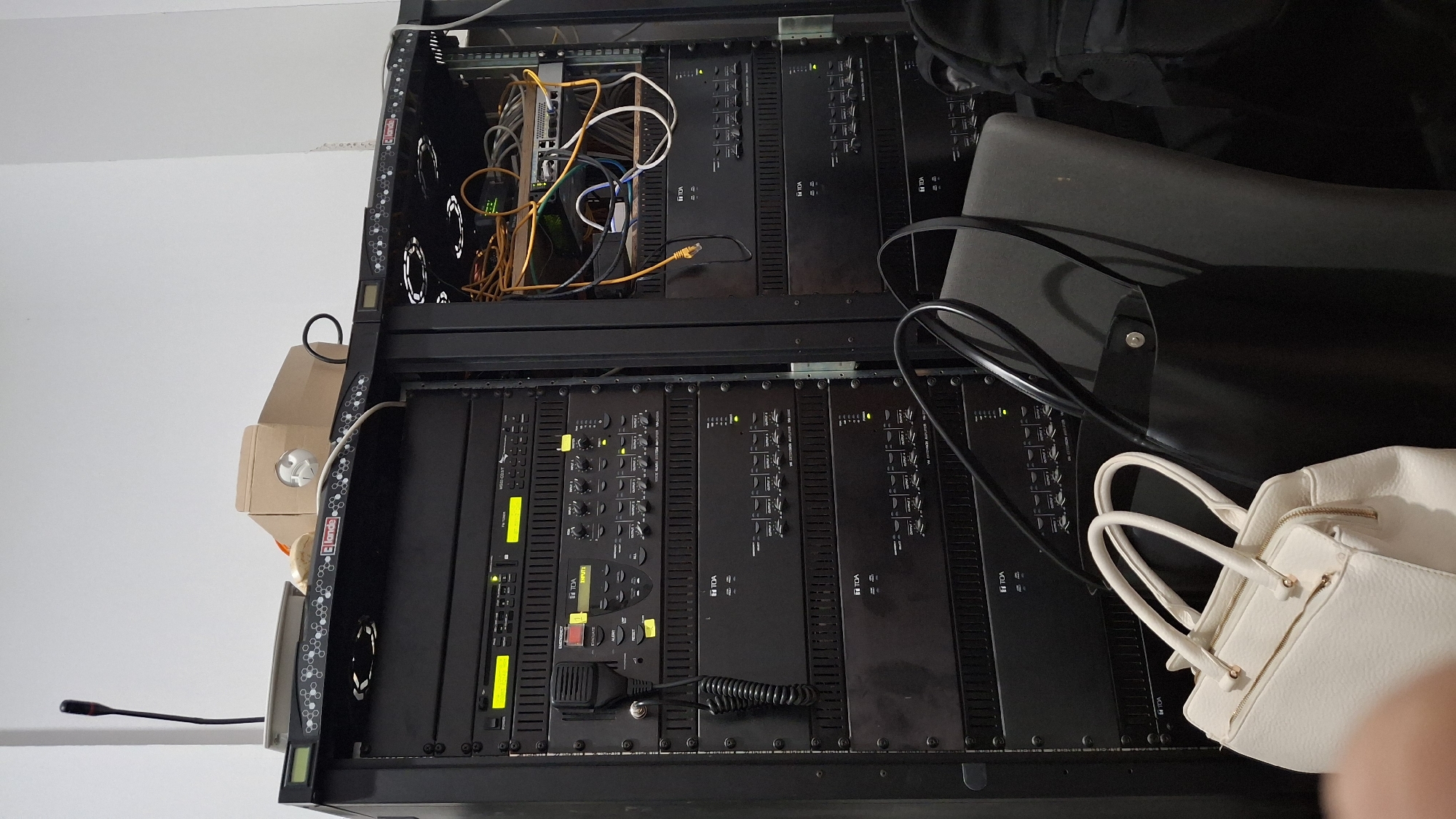 In Nesebar, Bulgaria, this image captures a rack of electronic audio equipment. The rack contains multiple black units, including amplifiers and audio system components, some bearing the "TOA" logo. In the foreground, two handbags are visible: a white textured handbag on the left and a black tote bag on the right, resting on a grey chair. Cables, predominantly orange and blue, are visible in the upper right section of the rack, connected to a network switch. A microphone with a coiled cord is attached to one of the audio units. The overall environment suggests a technical setup, possibly in a venue or control room.
