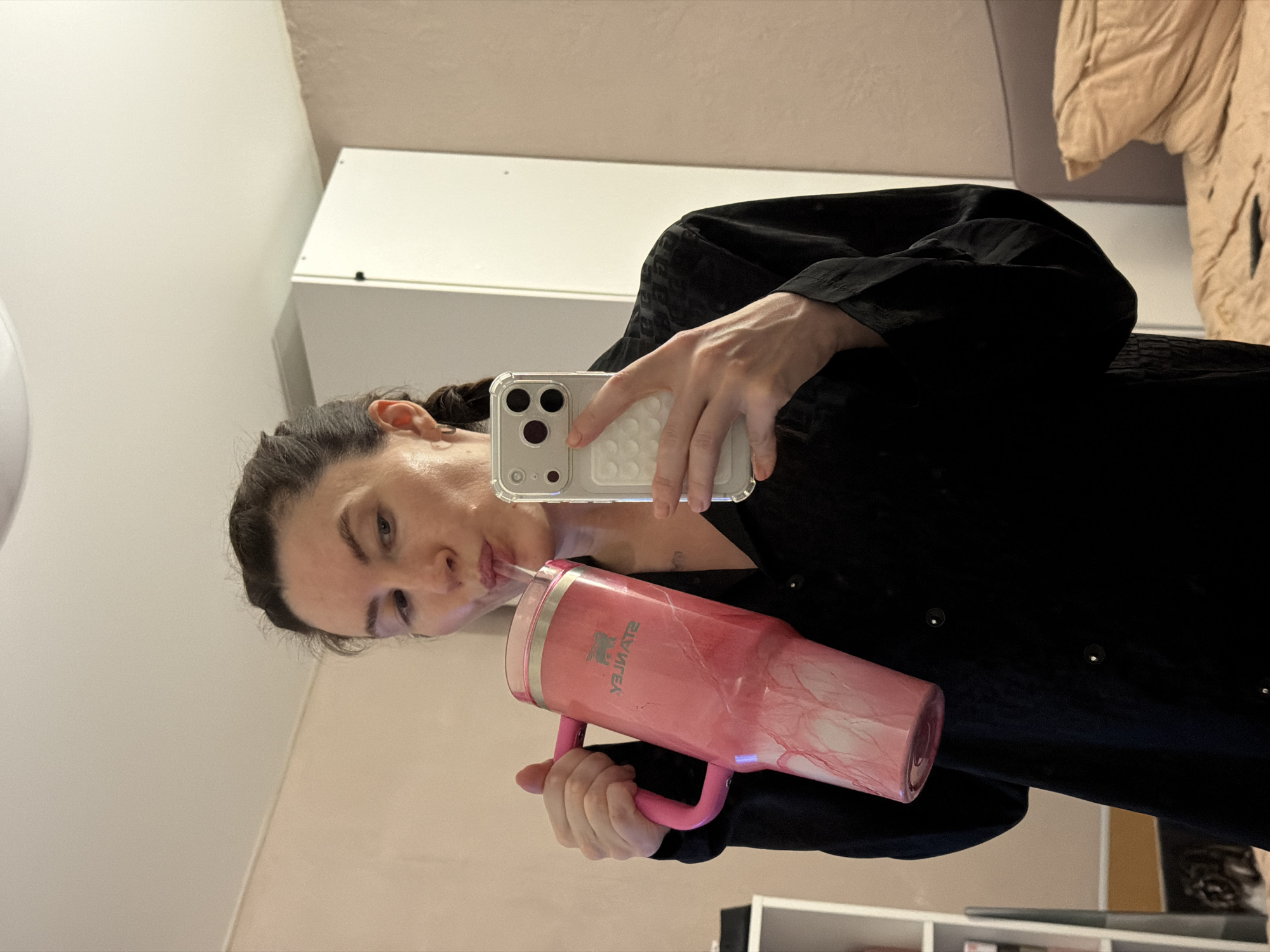 A female individual is depicted in a mirror selfie taken within an interior room located in Perm, Russia. She has dark hair styled in a braid and wears a dark, possibly patterned, long-sleeved shirt. Her left hand holds a large, pink Stanley tumbler with a marbled design, a pink handle, and a clear straw, from which she is actively drinking. Her right hand holds a light-colored smartphone in a clear case, featuring a visible triple-lens camera array, used to capture the image. The background shows a light-colored wall, a partial white cabinet or wardrobe unit to her left, and a bed with a brown headboard and crumpled beige bedding to her right. A white shelving unit is partially visible behind her left side.
