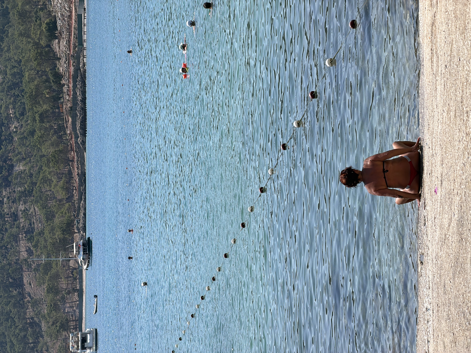 A woman in a red bikini sits on a pebbly beach facing the clear, blue water of the Mediterranean Sea in Kemer, Turkey. In the distance, sailboats and yachts are moored near a tree-covered hillside. A line of buoys separates the swimming area from the open water. Several people are visible swimming or wading in the sea. The scene captures a tranquil summer day at the beach.