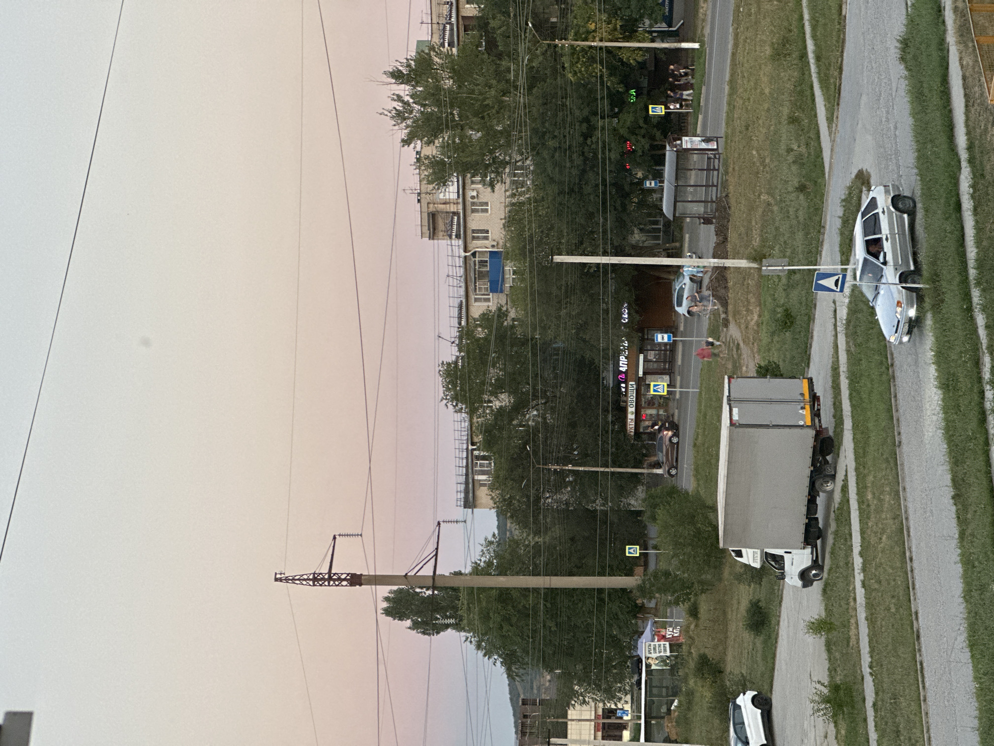 The image depicts an urban scene at dusk in Nevinnomyssk, Russia, characterized by a soft, pinkish sky. In the foreground, a silver sedan is navigating a road intersection. To the left, a white box truck is parked on asphalt. The middle ground features a road with a bus stop, several pedestrians, and a few cars, including a brown sedan. Trees and residential buildings line the street, with power lines crisscrossing the sky and a prominent utility pole with a lattice-top structure on the left. The overall environment suggests a typical neighborhood in the city.