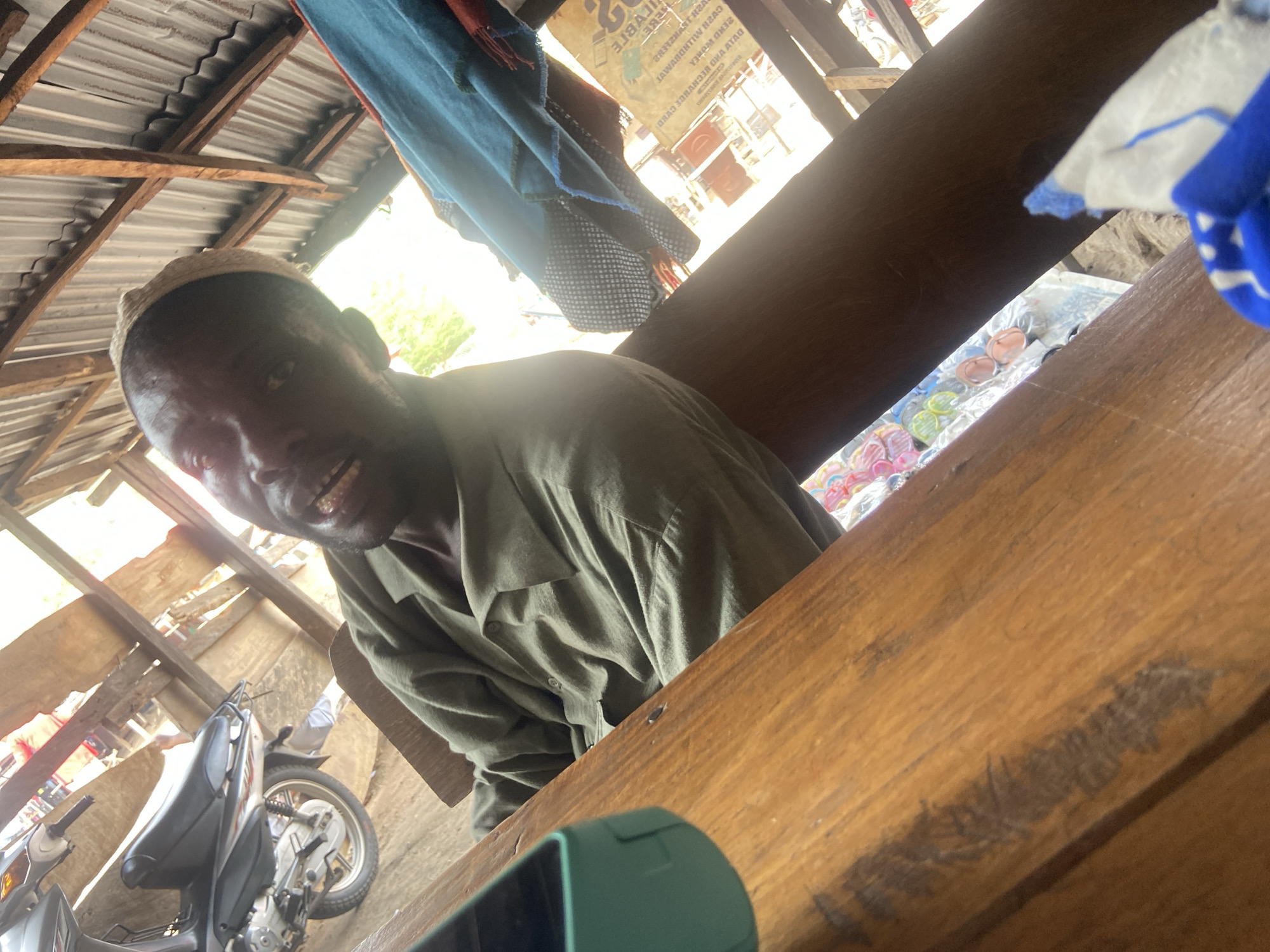 The image shows a man with dark skin wearing a light-colored cap and an olive green collared shirt, sitting at a wooden table. The table is in the foreground, taking up a significant portion of the image. In the background, visible through the gaps in the structure, there is a motorcycle parked outside, suggesting a possible outdoor or semi-outdoor location. Overhead, there's a corrugated metal roof. Part of a blue fabric item hangs from the ceiling. The scene appears to be in a bustling environment, indicated by the presence of various objects and structures in the background. It is difficult to determine the location as Mararaba, Nigeria, solely based on the visual information provided, but the overall scene suggests a somewhat informal or perhaps even a roadside setting. The man is looking at the camera with an apparent smile. The image appears to have been taken indoors, capturing the subject in what seems to be a candid moment.