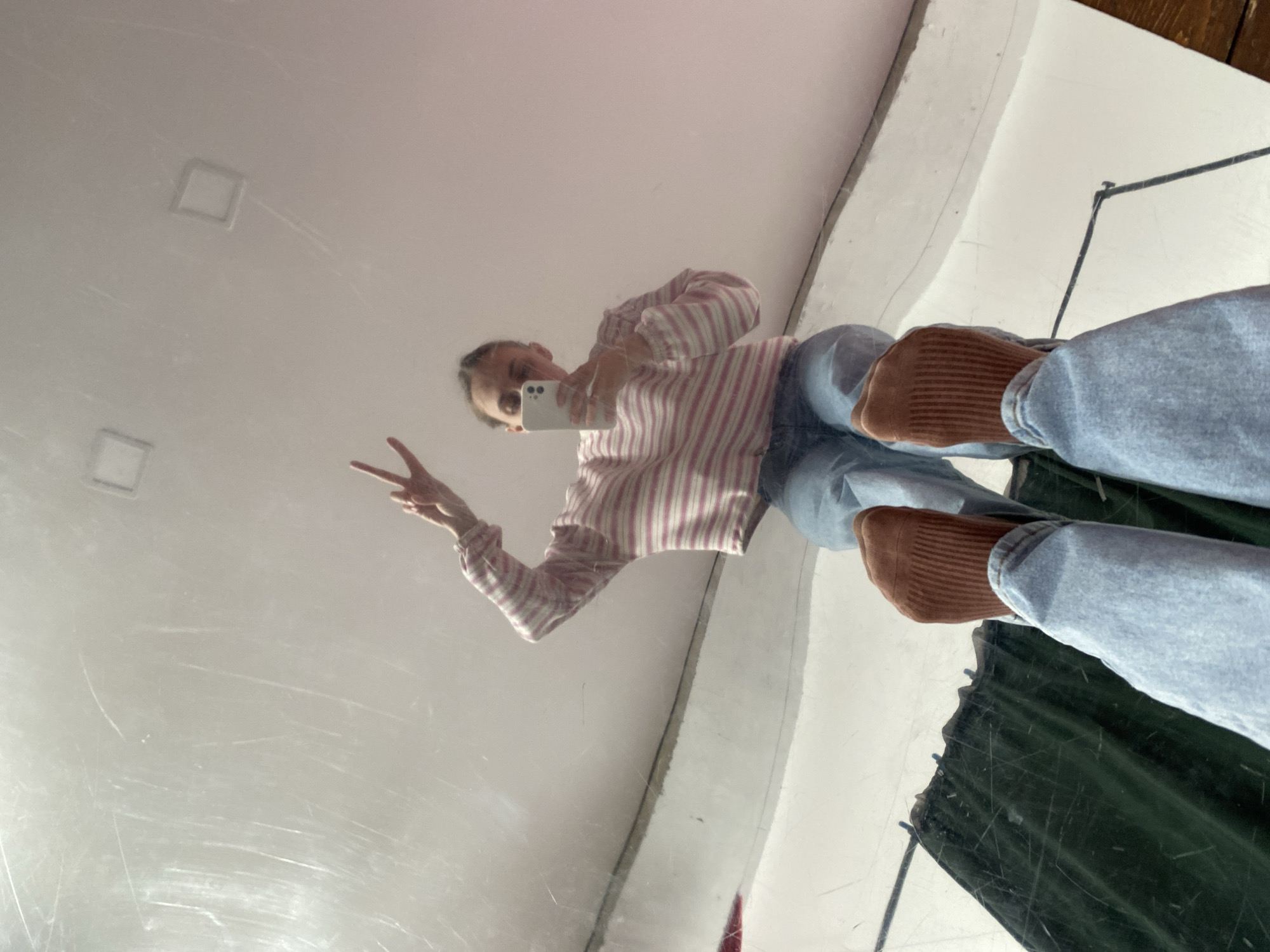 A young woman is reflected in a large mirror. She is holding a white smartphone, possibly taking a selfie, and making a peace sign with her left hand. The woman is wearing a pink and white striped shirt, light blue jeans, and brown socks. The mirror's surrounding environment includes a white ceiling with square light fixtures, a white wall, a dark green curtain, and a portion of a wooden surface. There is no clear evidence to confirm the exact location within Chaikovsky, Russia.