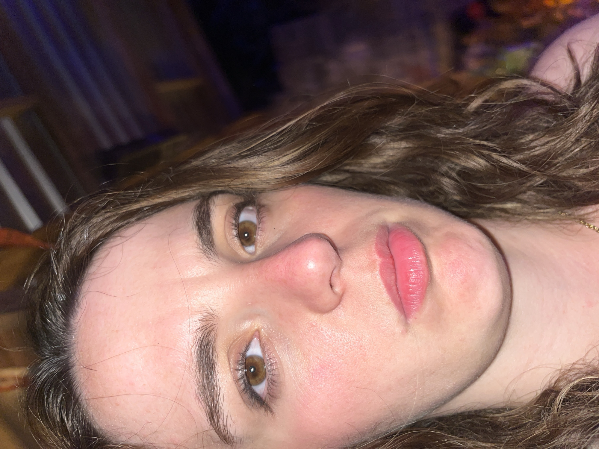 A close-up, slightly angled, indoor shot shows a young woman's face. She has brown hair, light skin, and brown eyes. Her lips are pink and appear to have lip gloss or lipstick on them. There is a slight red coloration on her cheeks and nose. The background is dark and indistinct, with some blurred lights visible. No specific location information is discernible from the image.