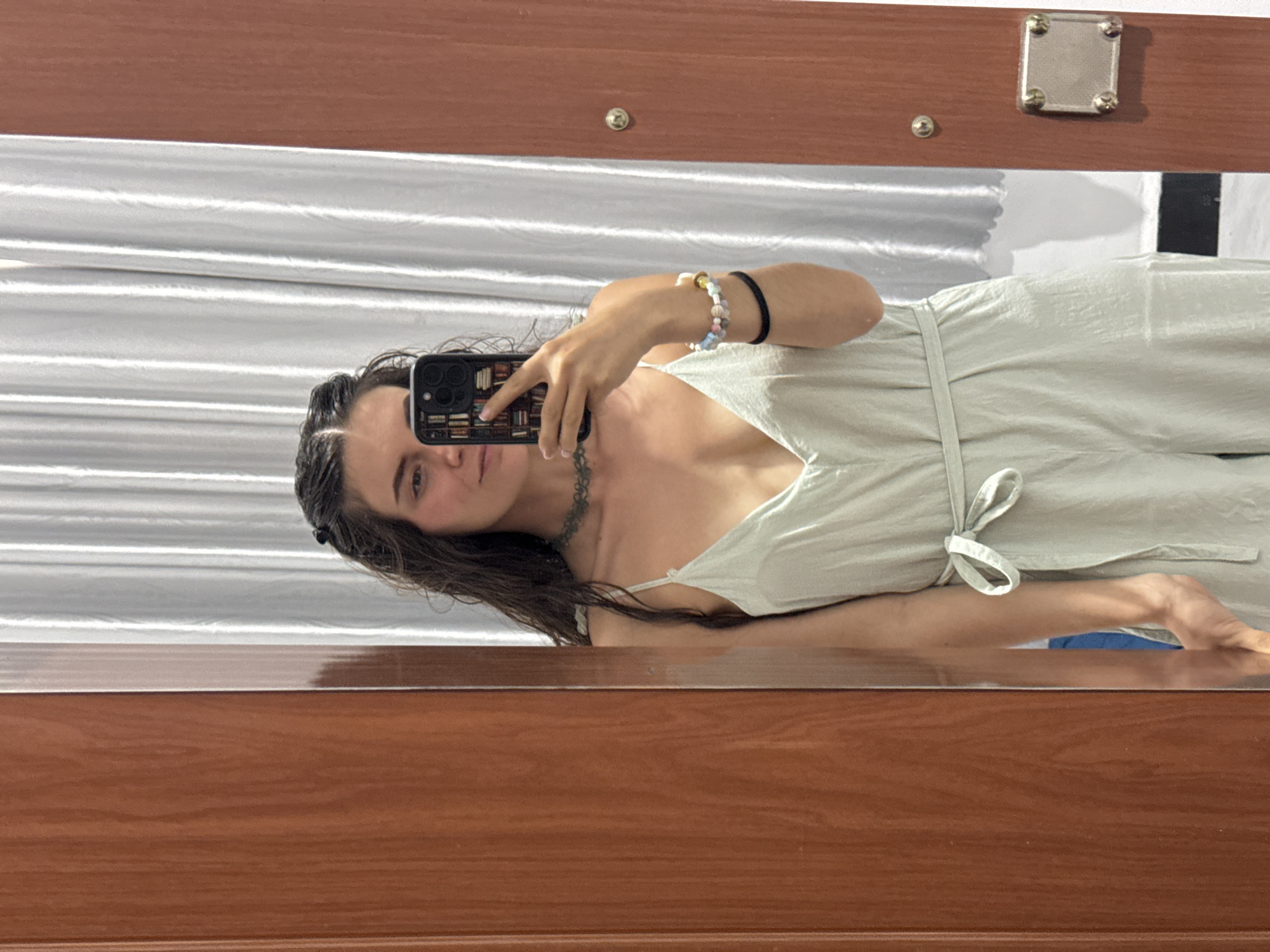 The image depicts a young woman taking a selfie in a mirror. The woman is light-skinned with dark, wet hair. She is wearing a light green jumpsuit with a tied waist. She is holding a smartphone with a dark case in her right hand, obscuring part of her face. She is wearing a beaded bracelet and a black wristband on her right wrist. A delicate necklace is visible around her neck.
The background is a white curtain with horizontal folds. The mirror is framed by a brown wooden structure with a metal fixture in the upper right corner. The image is taken at an angle, with the subject appearing to be leaning slightly. The location, Mũi Né, Vietnam, cannot be directly determined from the visible elements in the image.
