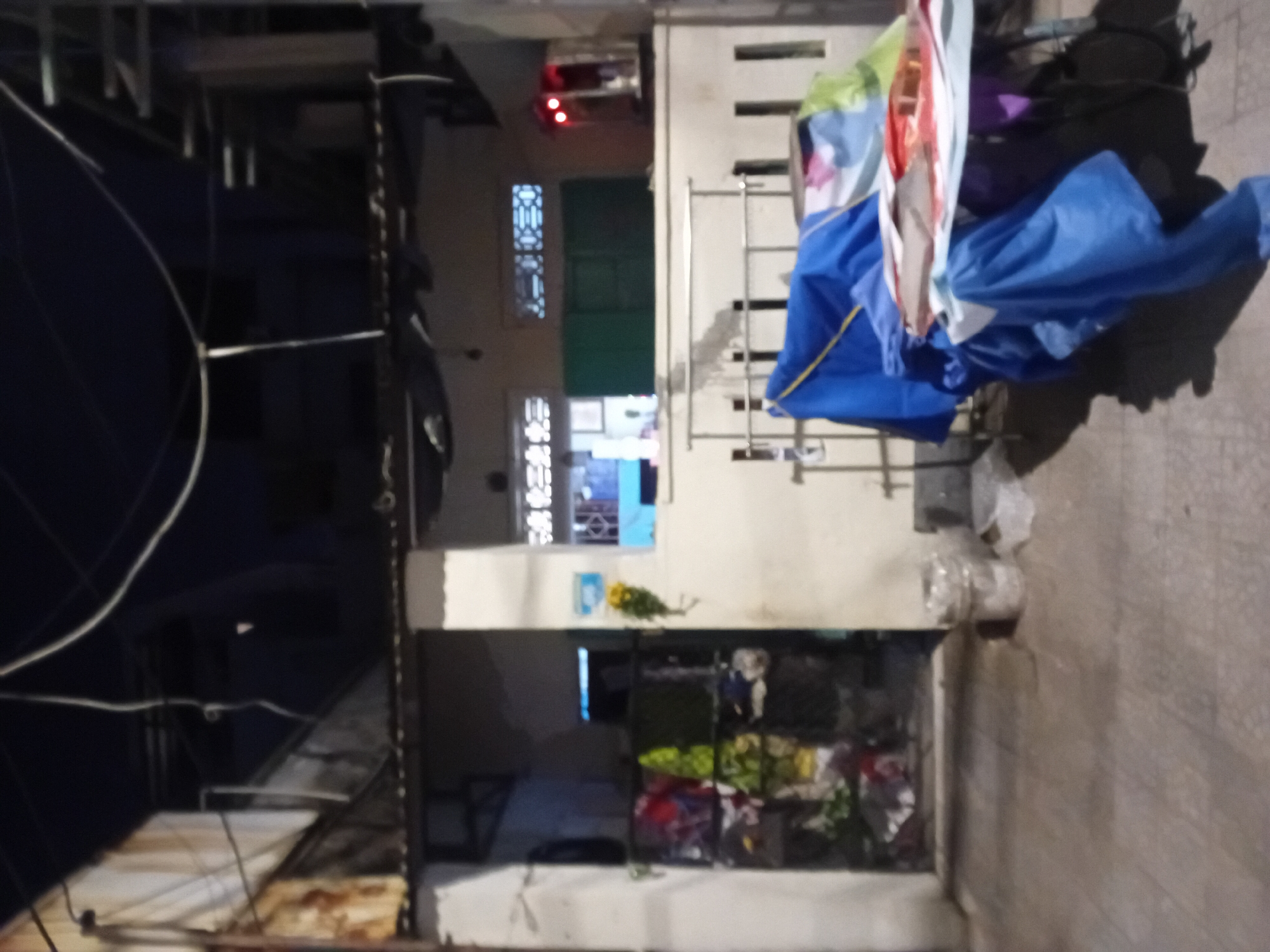 The image shows an exterior view of a building, likely a residential or commercial property, at night. The scene is dimly lit, suggesting it is dark outside, and the lighting appears to be artificial.
In the foreground, there is a table covered with various items, including blue tarpaulins, papers, and possibly some fabric or clothing. A bicycle is partially visible beneath the table. The ground is paved with what appears to be stone or concrete tiles.
The middle ground features a building facade. To the left, a dark opening, possibly a doorway or gate, is visible. Inside this opening, laundry is hanging, with various colors and patterns of clothing apparent. Adjacent to this opening, a white pillar stands, and a yellow flower is attached to it or hanging nearby. To the right of the pillar, a window with an intricate decorative grille is visible, and beyond it, another window is partially obscured by a blue element. Further to the right, a green structure, possibly a door or shutter, is present. Above the green structure, two rectangular windows with decorative patterns are visible. On the far right of the middle ground, a small structure with red lights illuminated is present, resembling a street food stall or an illuminated sign.
The background shows the upper parts of buildings and structures, with electrical wires crisscrossing overhead. The sky is dark.
The scene suggests a modest urban environment. The presence of laundry hanging outside, the somewhat cluttered appearance of the table, and the general state of the building might indicate a working-class neighborhood. The time of day (night) contributes to the overall atmosphere of the scene.
Based on the information provided in the prompt, this scene is located in the city of Đà Nẵng, Vietnam. The architecture, the style of windows, and the presence of laundry hanging outside are consistent with observations from urban areas in Vietnam. The image captures a moment in the daily life of this location.