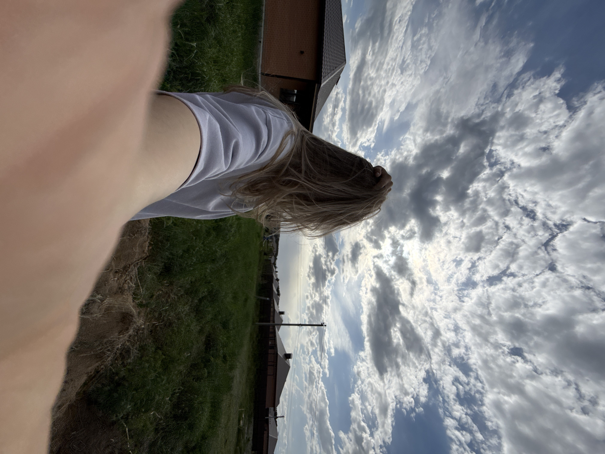 A distorted, low-angle selfie perspective captures a person lying on the ground, with the sky filling most of the frame. The sky is a mix of blue and white, with numerous puffy clouds scattered across it. The person has blonde hair and is wearing a light-colored t-shirt. A portion of grass and what appears to be the edge of a dirt patch are visible on the left side of the image. In the background, several buildings with brown roofs and a utility pole can be seen. The scene appears to be located in Chaikovsky, Russia, evidenced by the architecture and typical landscape.