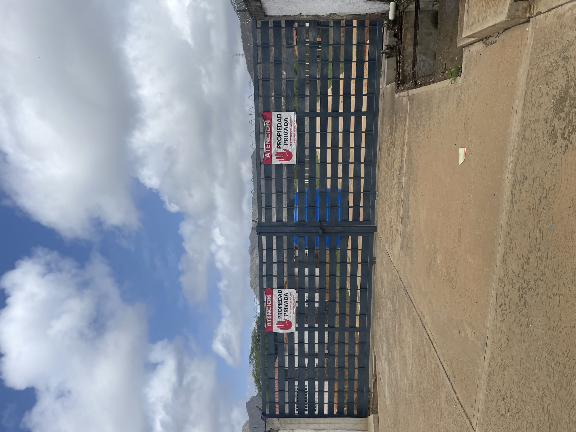 The image shows a closed, dark gray metal gate with a barbed wire fence on top. Two red and white signs are affixed to the gate. The sign on the left reads "ATENCION PROPIEDAD PRIVADA SE RESTRINGE EL ACCESO SIN AUTORIZACIÓN". The sign on the right is identical. A blue trash receptacle is visible behind the gate. The gate is set against a backdrop of a concrete wall on the left and a more rustic wall on the right, with a partial view of buildings and foliage behind the gate. The ground in front of the gate is concrete. The sky is cloudy with patches of blue. This scene is located in El Valle del Espíritu Santo, Venezuela.