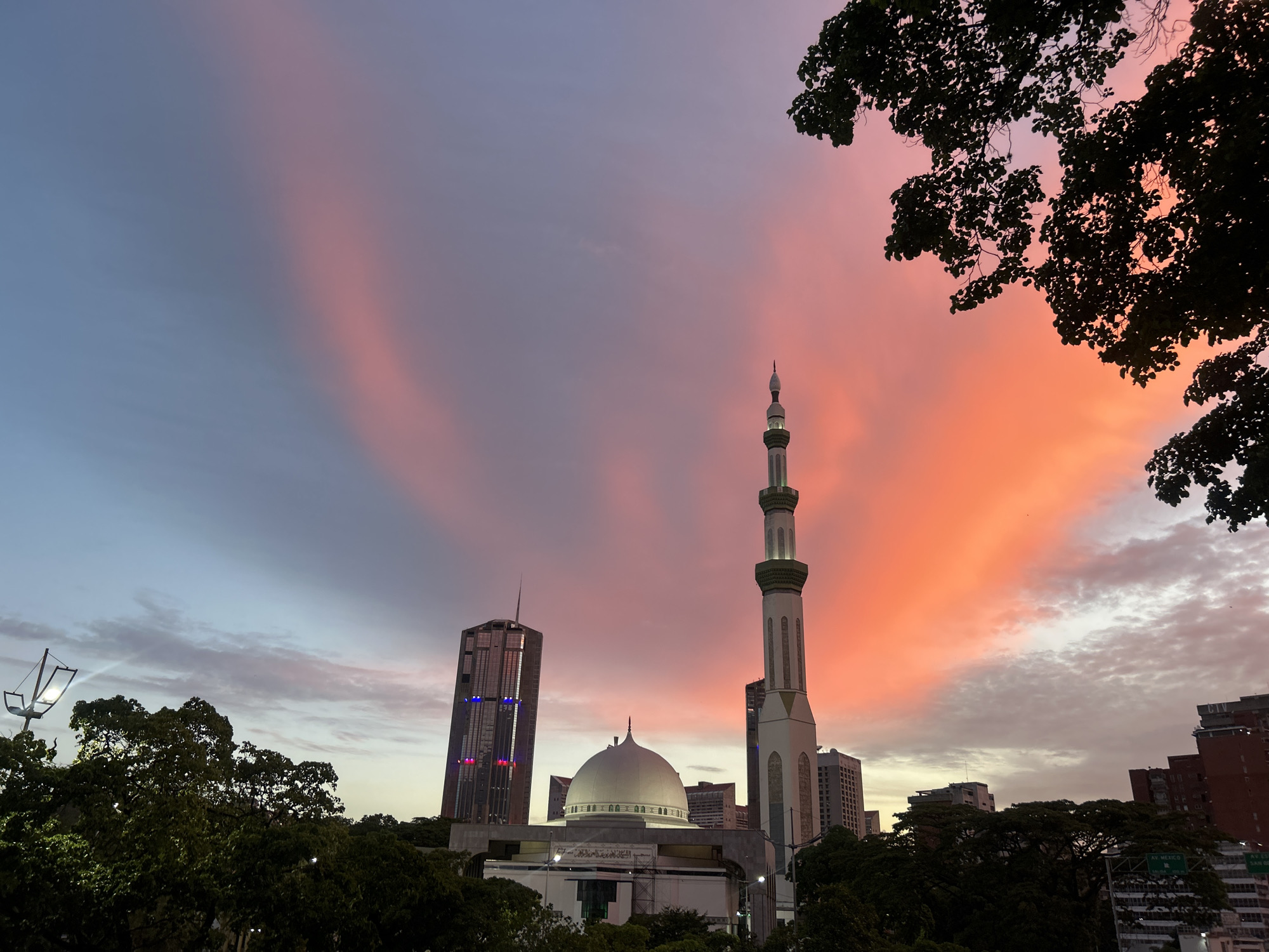 The image shows a mosque in Caracas, Venezuela, at dusk or dawn. The sky is a gradient of blues and pinks/oranges. A tall minaret rises from the mosque, and a large dome is visible. A modern skyscraper stands to the left of the mosque. Trees frame the bottom and right edges of the image. The mosque is surrounded by greenery. Other buildings are also visible in the background.