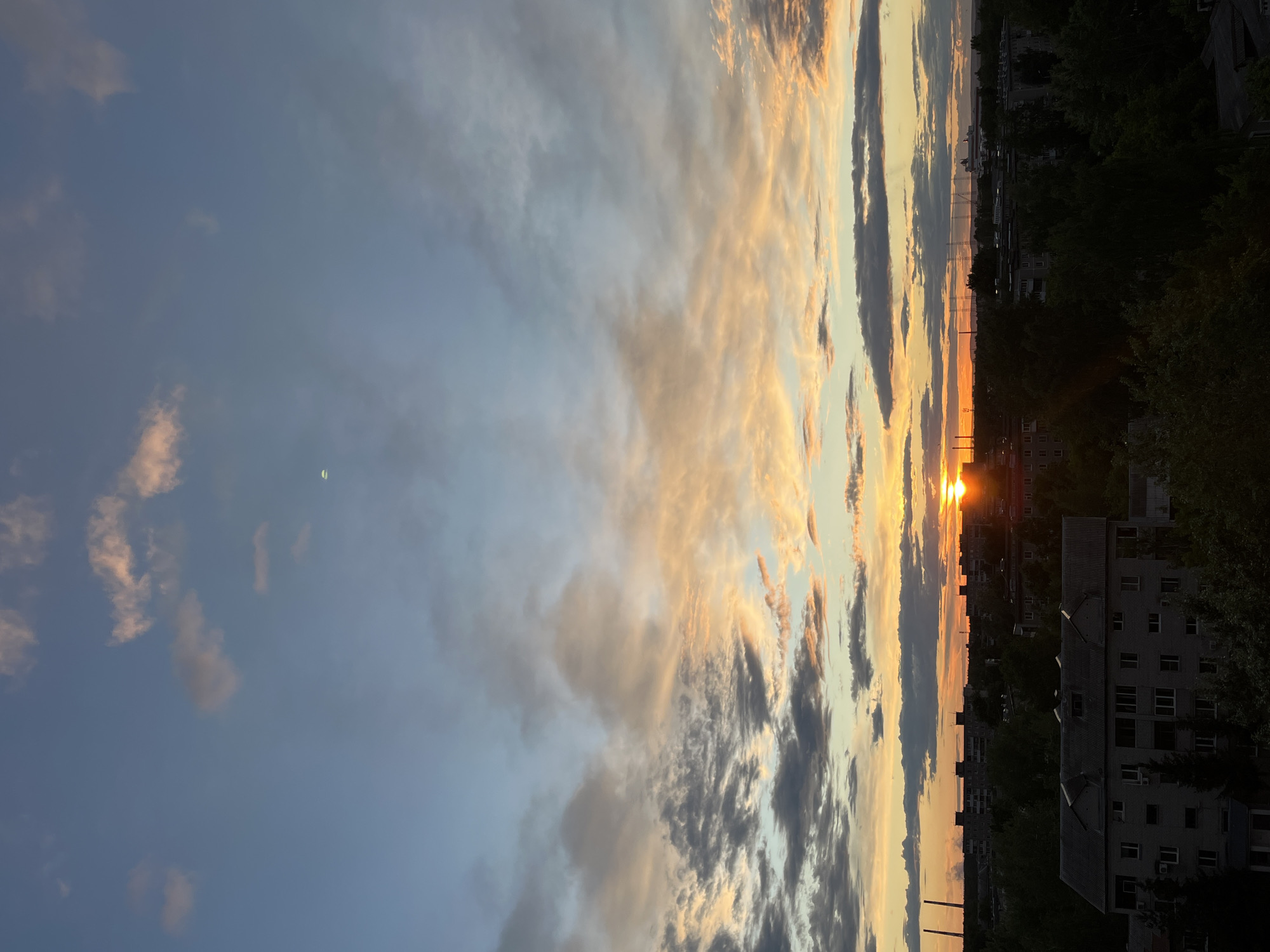 The image depicts a sunset over an urban area. The sky is filled with clouds, with hues of blue, gray, and orange from the setting sun. Below the horizon, a cityscape is visible, featuring buildings and trees. The overall scene suggests the image was taken in an urban environment, specifically in the city of Unknown, Unknown. The buildings are multistory and densely packed. The photograph appears to have been taken from an elevated position, offering a wide view of the city.
