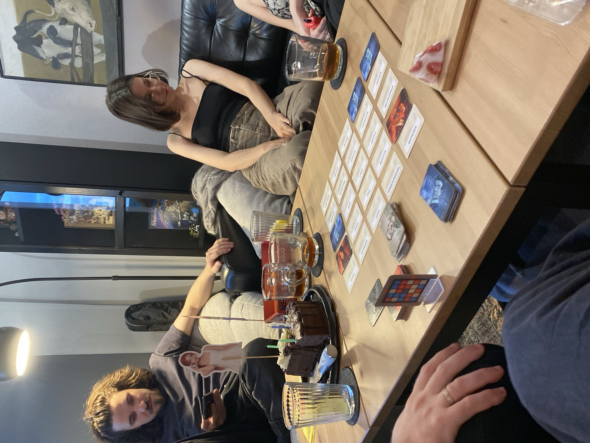 People are sitting around a wooden table, playing a card game. There are playing cards spread across the table and several glasses of alcoholic beverages, along with a piece of chocolate cake. There are two women and one man visible in the image. The environment appears to be an indoor space, possibly a home or a cafe, with a painting of cows hanging on the wall. It is not possible to confirm whether this is the city of Moscow, Russia, based on this single image.