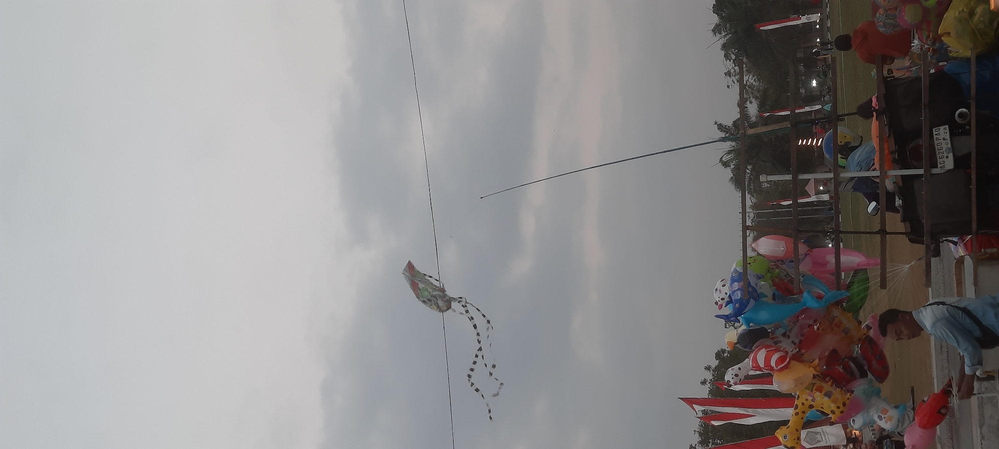 In Kanigoro, Indonesia, on an overcast day, a kite with a patterned design and long streamers flies in the sky, tethered to a wire. Below, a collection of colorful balloons, including animal shapes like a giraffe and a dolphin, are displayed on a cart. A man in a light blue shirt is seen attending to the balloons, while other people are visible in the background. Red and white flags, likely representing the Indonesian flag, are also present. The scene appears to be a public gathering or event, possibly a market or festival.