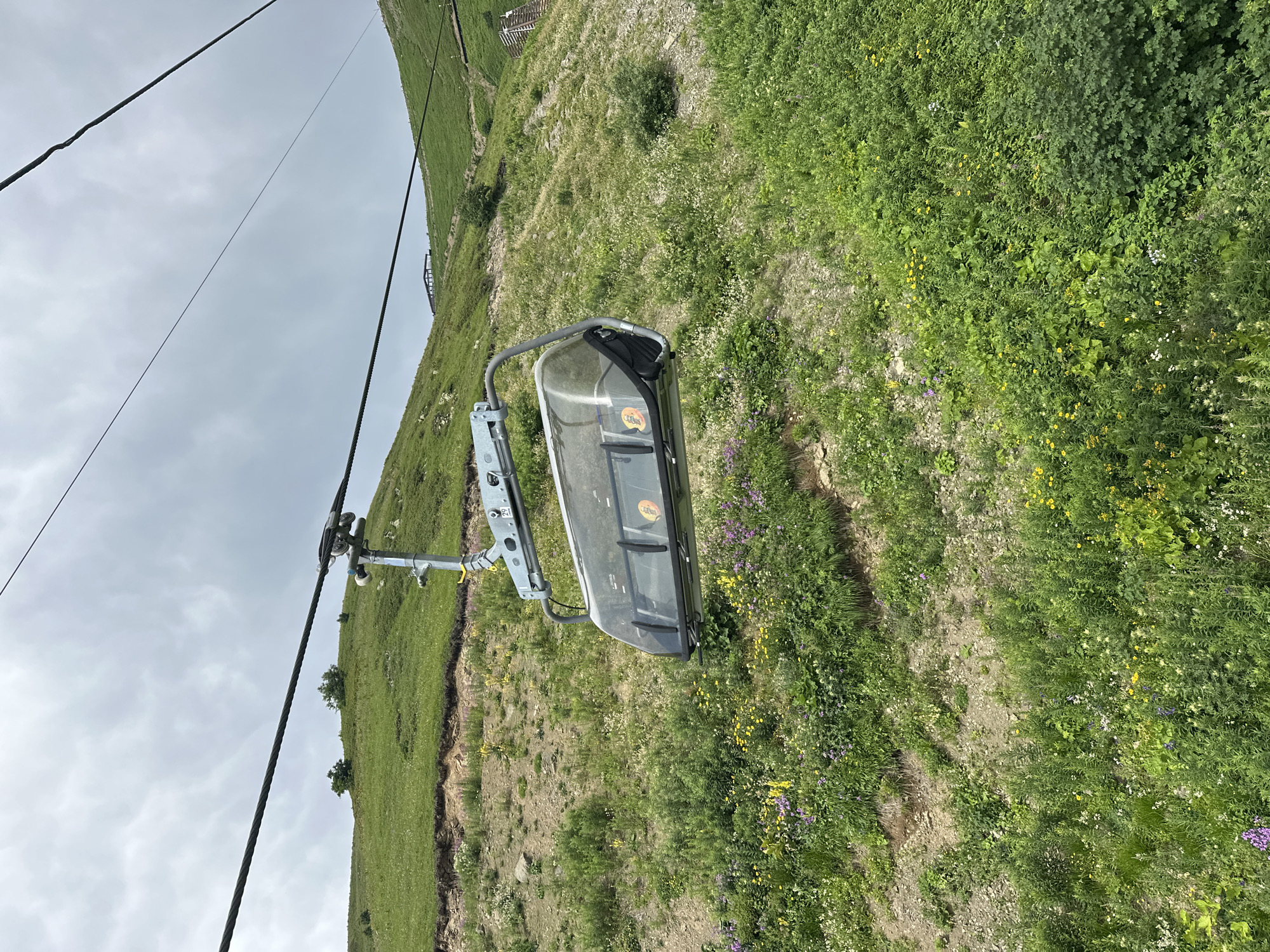 The image shows a ski lift car suspended on a cable over a steep, grassy slope in Estosadok, Russia. The slope is covered in lush green vegetation and wildflowers of various colors, including yellow, purple, and white. The sky is overcast with gray clouds. The ski lift car is transparent, revealing empty seats inside. The cable runs diagonally across the frame, disappearing into the upper right corner. The scene depicts a mountain landscape during the warmer months, likely summer or early autumn, when the ski slopes are not covered in snow.