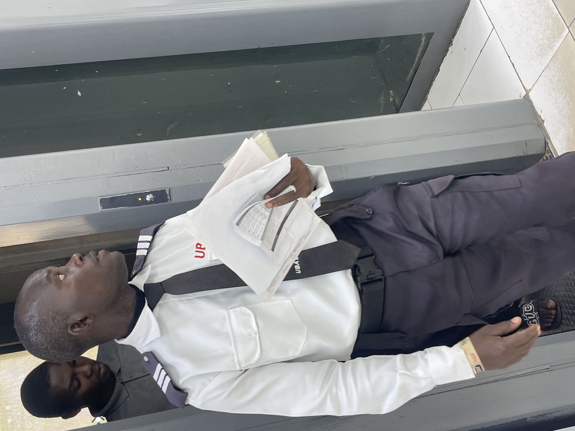 An indoor scene in Zaria, Nigeria, shows a man in a security guard uniform lying on what appears to be a conveyer belt, perhaps at a checkpoint or scanner location. He is wearing a white, long-sleeved shirt with "UP" in red lettering on the upper right chest, a black tie with "UBA" visible, and dark pants. He holds papers in one hand. A second, unidentified man is partially visible in the lower left corner of the frame. The floor appears to be tiled.