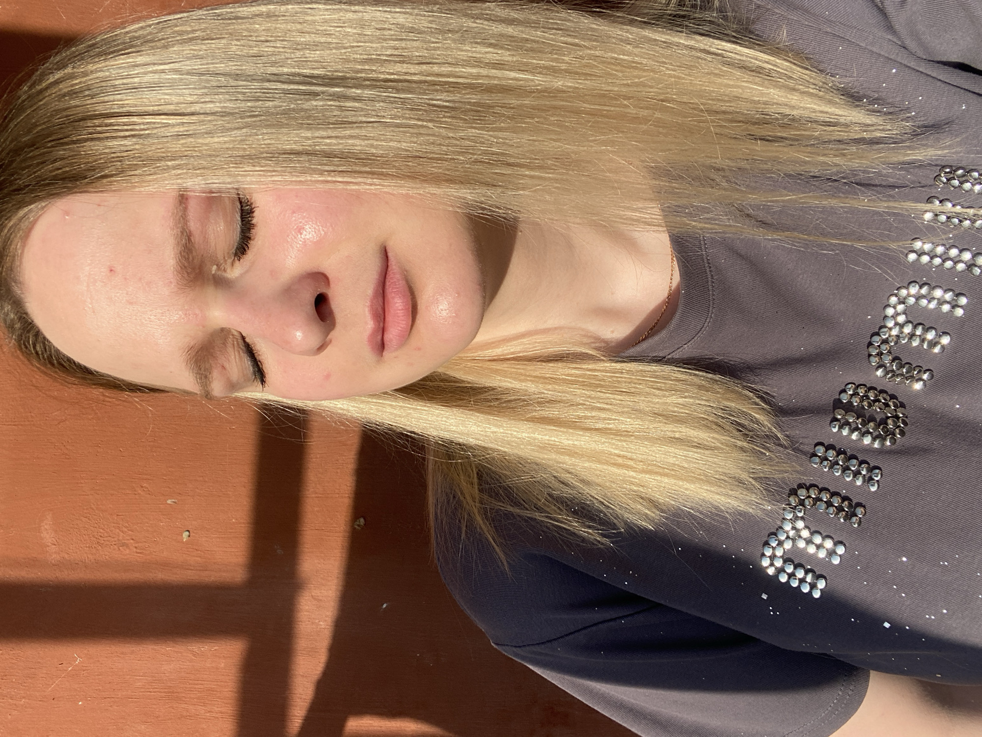 A young woman with light blonde hair and fair skin is shown in an outdoor setting, lying against a reddish-brown surface. Her eyes are closed, and she has a serene expression. She is wearing a gray t-shirt with what appears to be decorative silver studs or stones forming a word or design. A thin gold chain is visible around her neck. The setting appears to be sunny, with shadows cast across the wall behind her. The location in the city is listed as Unknown, Unknown.