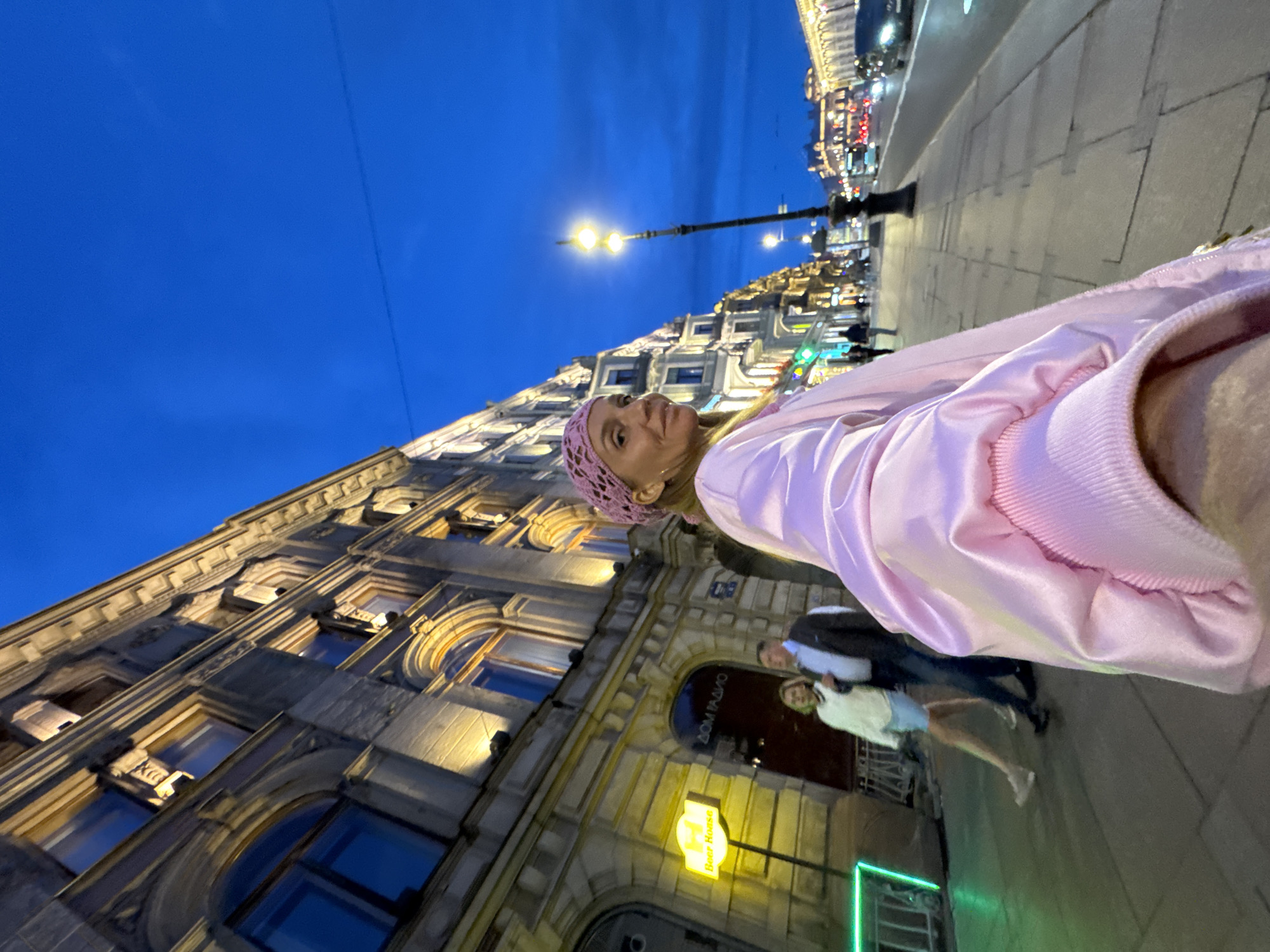 In Saint Petersburg, Russia, at dusk, a woman with blonde hair and a pink knitted headscarf is walking on a sidewalk, looking towards the camera. She is wearing a flowing pink satin jacket. In the background, a man and a woman in casual attire are walking away from the camera on the same sidewalk. A neon green sign reads "Beer House" and a sign in Cyrillic reads "ДОМ ГАДИО" on the building. The street is lined with ornate, illuminated buildings, and a street lamp is visible. Cars are driving on the road to the right, and the sky is a deep blue, indicating twilight.