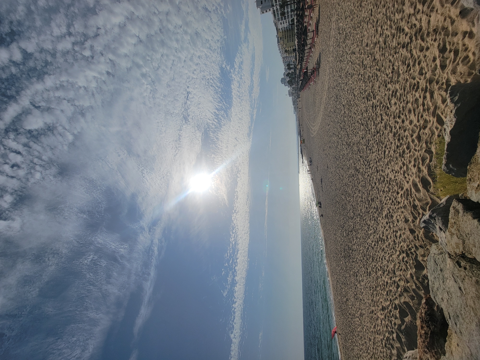 This media file depicts a wide, sandy beach with the ocean on one side and buildings on the other. The sun is shining brightly, with visible lens flare, and the sky is partly cloudy with scattered white clouds. The sand is textured with footprints and patterns, suggesting it has been walked on recently.
In the distance, along the edge of the beach, there are rows of beach umbrellas and lounge chairs, indicating a resort or tourist area. Several buildings, appearing to be hotels or apartment complexes, are visible behind the beach umbrellas. The ocean is calm with gentle waves lapping the shore. The water reflects the sunlight, creating a shimmering effect.
There are a few people visible on the beach and in the water, but they are small in the frame, suggesting the beach is not overly crowded. One person is wearing a red garment, standing near the water's edge. The time of day appears to be daytime, likely late morning or early afternoon, given the position of the sun and the quality of light. The weather seems pleasant, sunny with some clouds.
The image is oriented vertically, with the sky occupying the left half and the beach and ocean the right half. The overall mood is tranquil and suggestive of a holiday or vacation. The location is Quarteira, Portugal, a coastal town known for its beaches. There is no visible text in the media file.