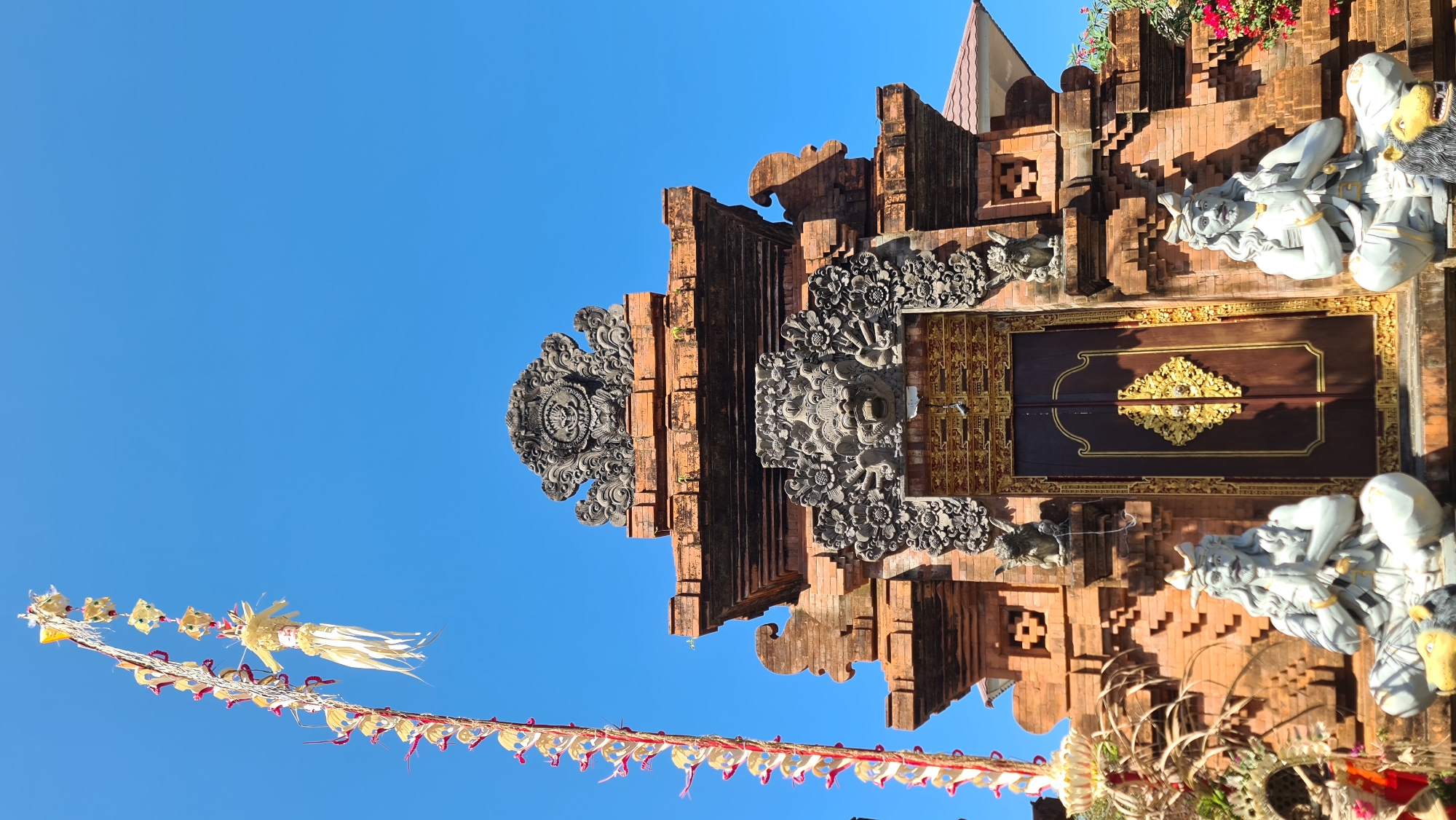 An exterior, eye-level, vertical, medium shot captures the ornate entrance of a traditional Balinese temple under a clear blue sky. The architecture is primarily constructed from reddish-brown brick, with intricate stone carvings adorning the gateway. A tall, decorative pole, adorned with yellow and red streamers and straw-like ornaments, stands prominently to the left of the frame.
Flanking the dark wooden double doors of the temple entrance are two seated stone statues. Both figures have long, wavy white hair and beards, wear ornate headwear, and are posed with their hands pressed together in a prayer-like gesture. They are seated with their legs crossed, and at the base of each statue, there is a depiction of a lion-like creature, one with a yellow face and the other with a black mane. The doors themselves are deep brown with gold embellishments, featuring a prominent diamond-shaped design in the center. Above the doors, a fearsome stone face with an open mouth and floral motifs surrounds it. The top of the gateway is topped with a decorative stone crest featuring a circular motif.
The temple appears to be located in Denpasar, Indonesia, a city known for its rich cultural heritage and numerous Hindu temples. The clear sky and bright sunlight suggest a warm, tropical climate. The overall scene conveys a sense of reverence and cultural significance, typical of religious sites in Bali.